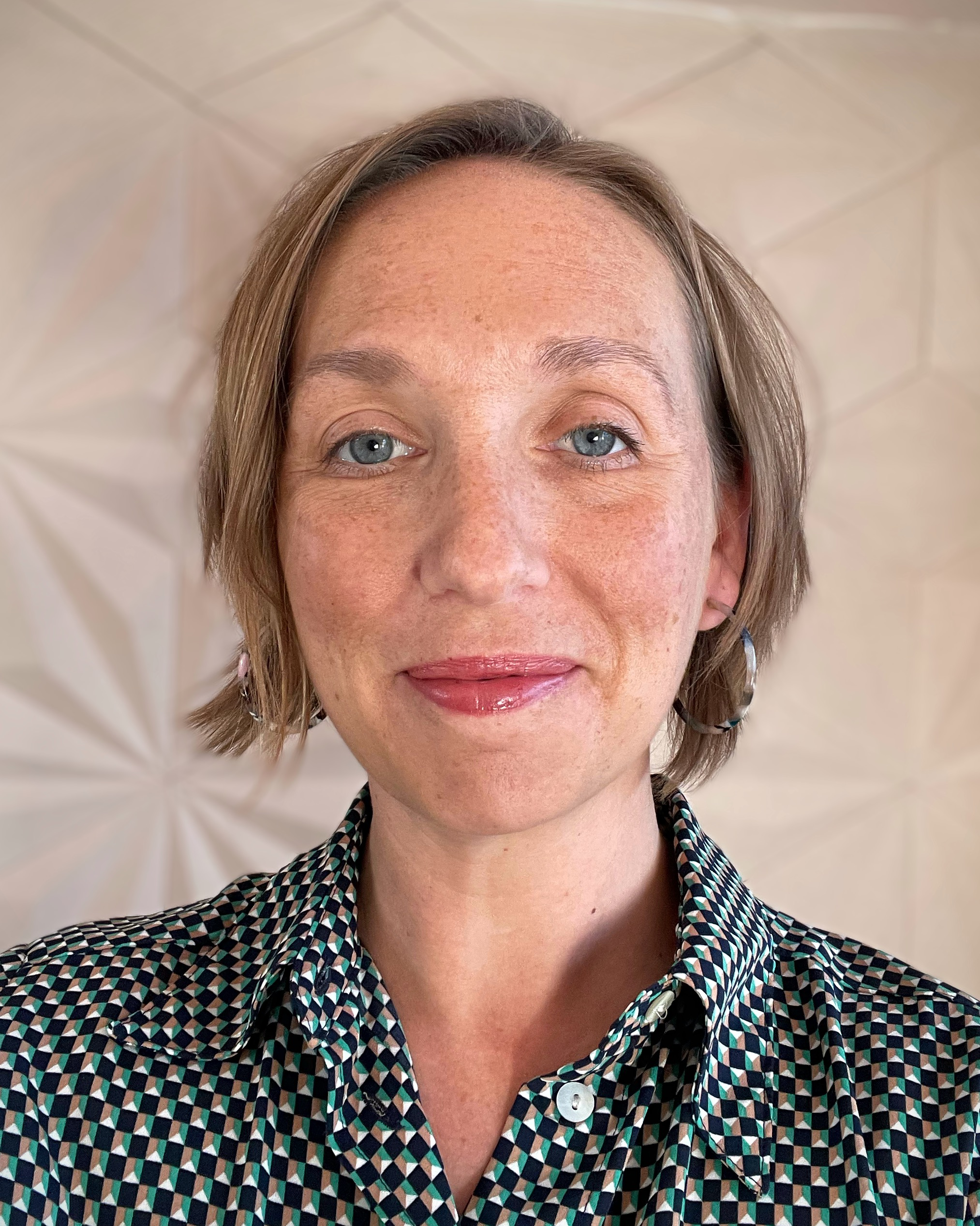 Close-up of a woman with short light brown hair, blue eyes, and fair skin with freckles, wearing a patterned shirt and earrings, smiling at the camera.