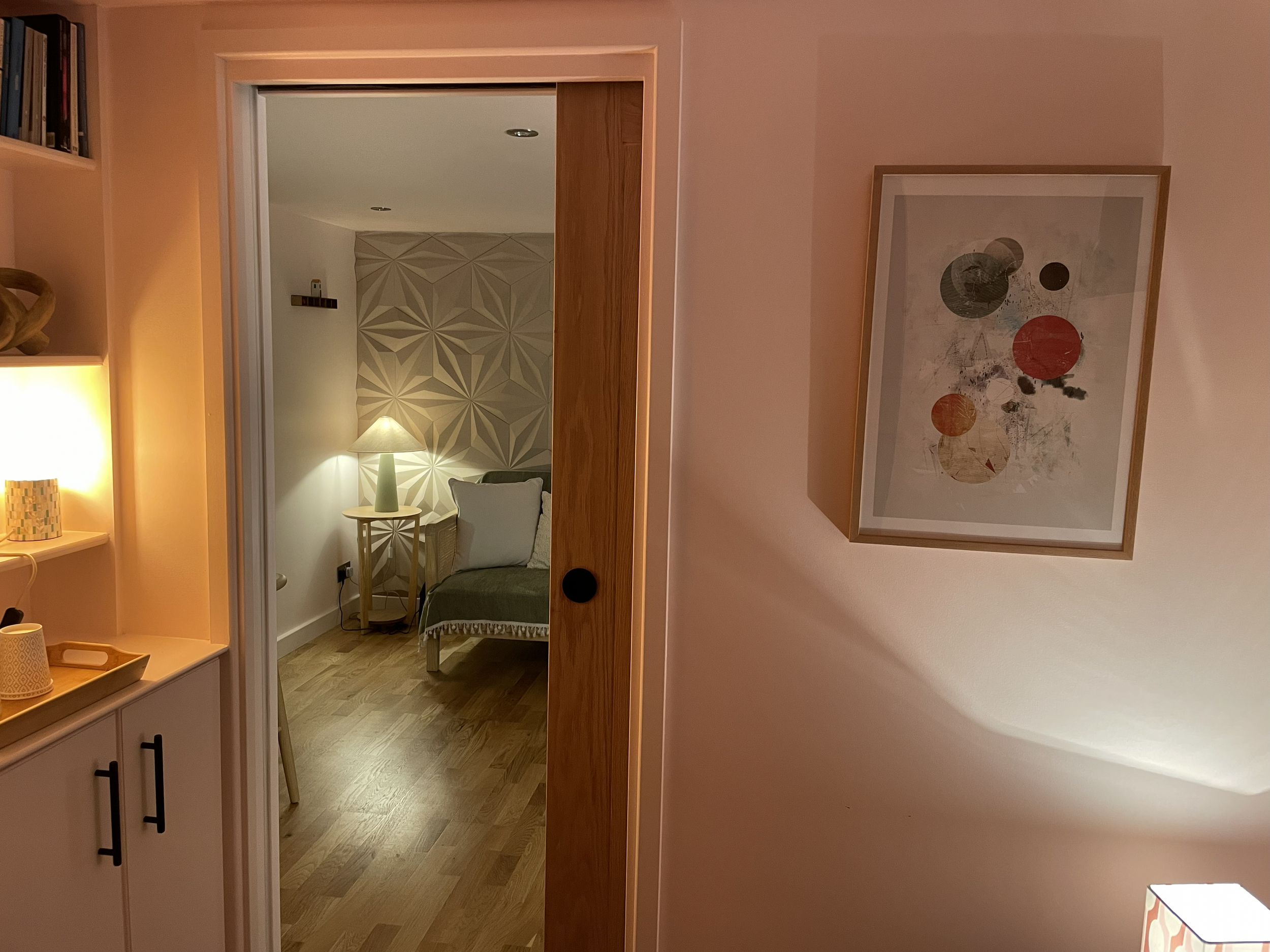 Interior view of a cozy living space with a bedroom visible through an open sliding door. The bedroom has a geometric accent wall, a bed with pillows, and a bedside table with a lamp. The foreground shows a wall with a framed abstract print and a cor