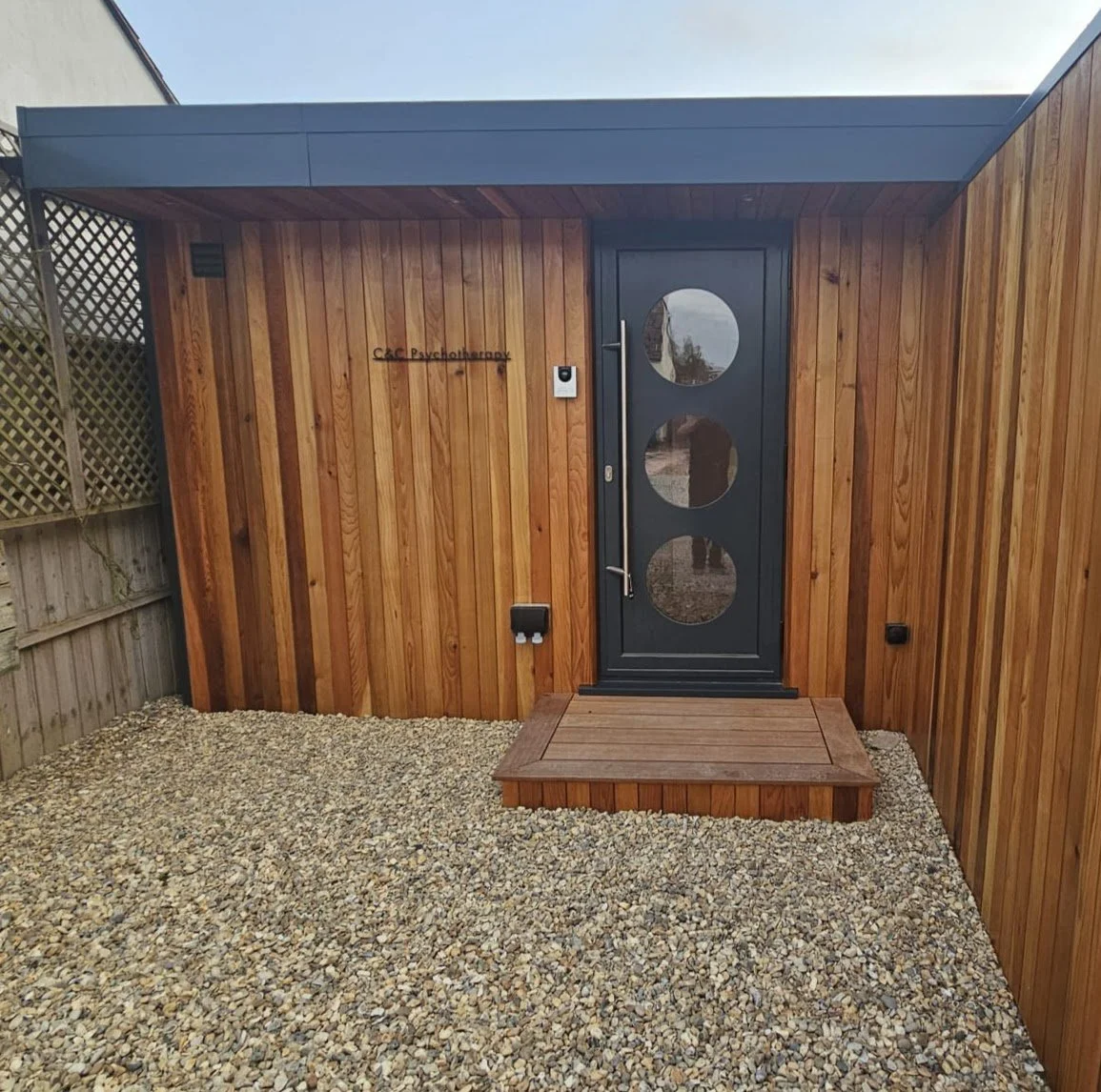 Wooden exterior of a psychotherapy clinic with a dark gray door with three circular windows, a small wooden platform in front, and gravel ground.