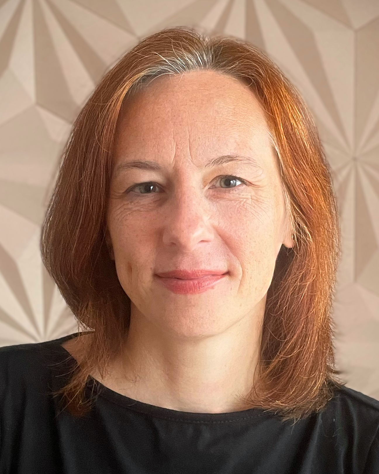 A woman with shoulder-length red hair, fair skin, and blue eyes, smiling slightly, wearing a black top, with a geometric-patterned background.