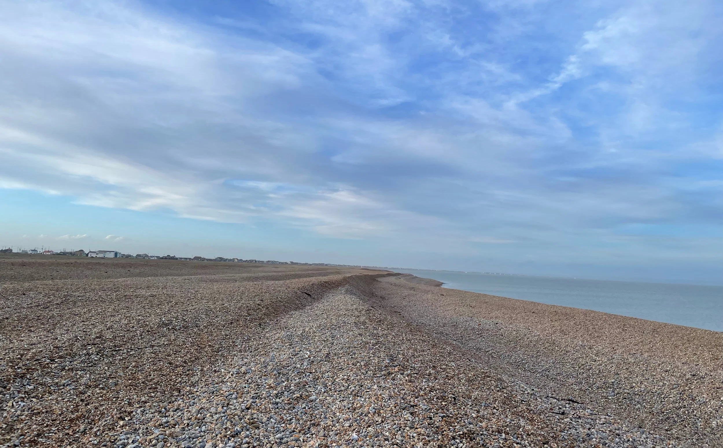 A pebbled beach stretches along the shoreline under a partly cloudy blue sky, with distant buildings visible on the horizon.