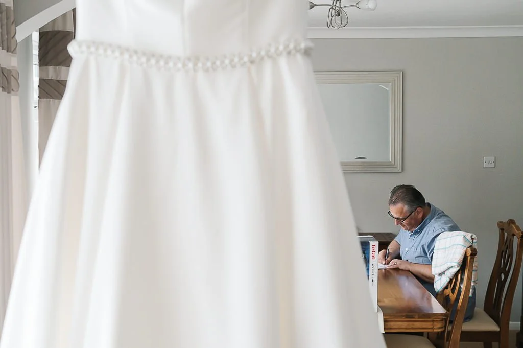 Brides father writing his speech with the wedding dress hanging in the foreground