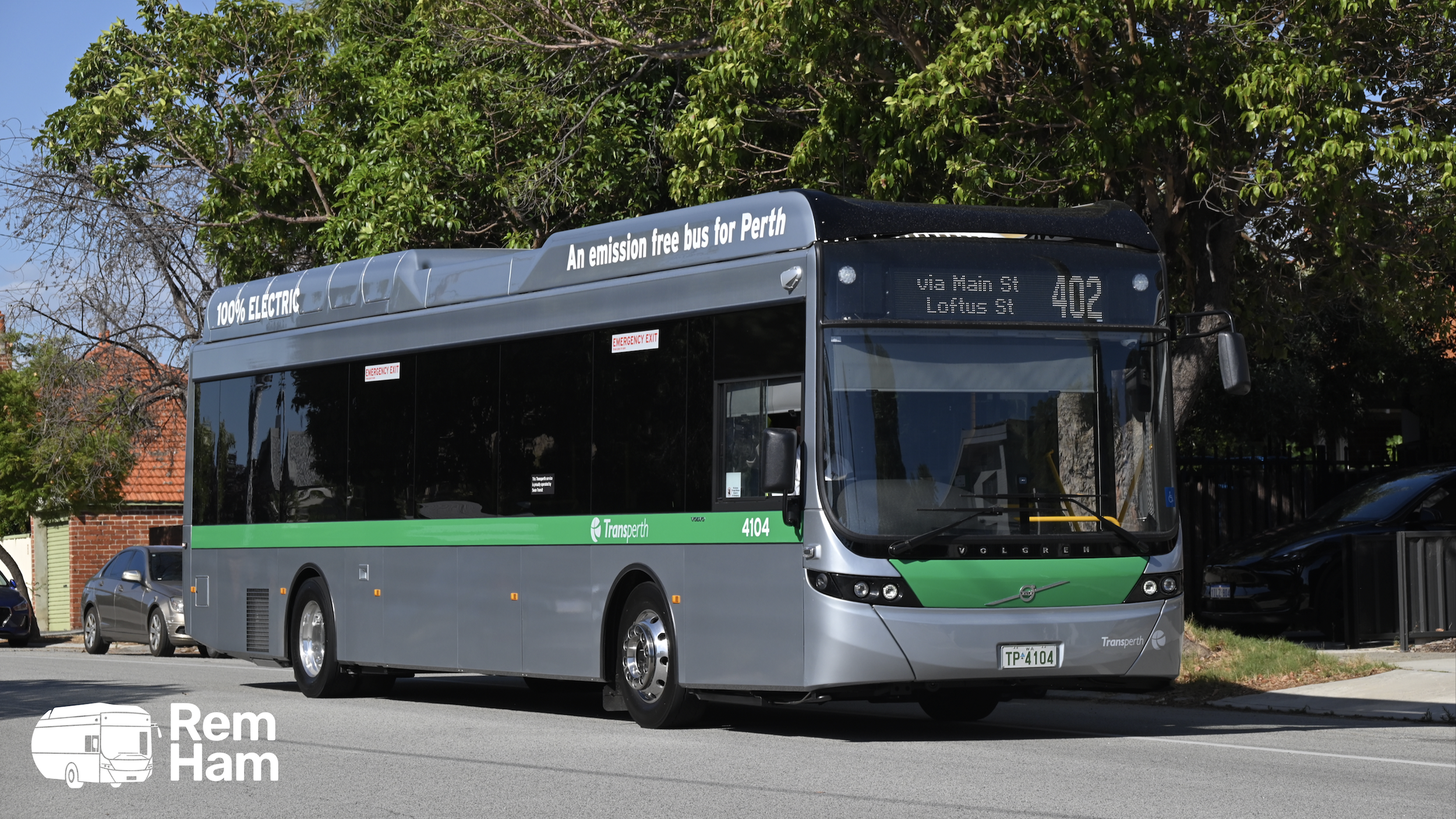 A green and gray electric bus with a sign indicating it is a zero-emission bus for Perth, displaying route 402 via Main Street and Lotus Street, parked on a street with trees and parked cars in the background.