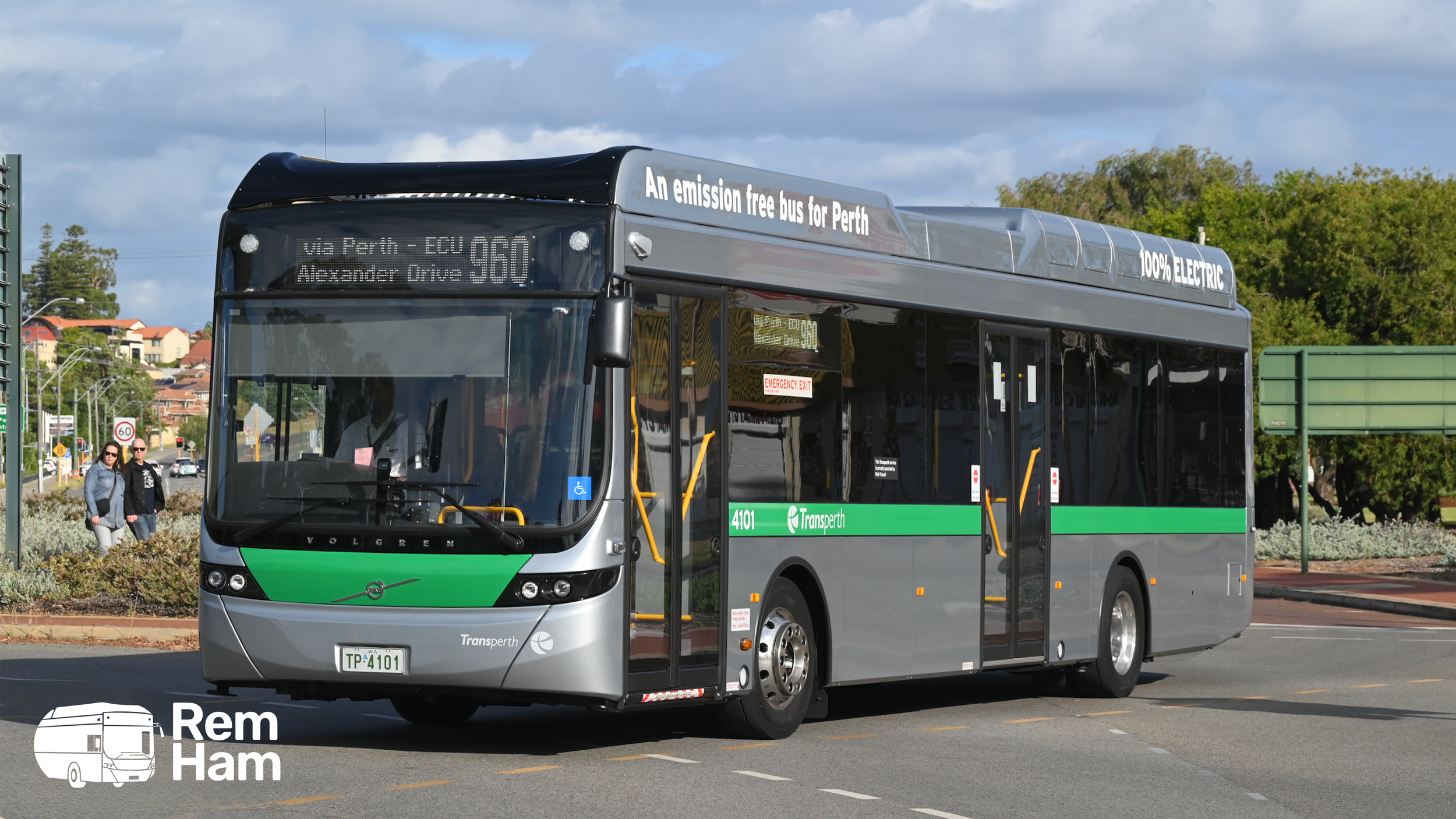 A gray and green electric bus on a city street with trees and buildings in the background, displaying signs indicating it is emission free and 100% electric, operated by TransPerth.