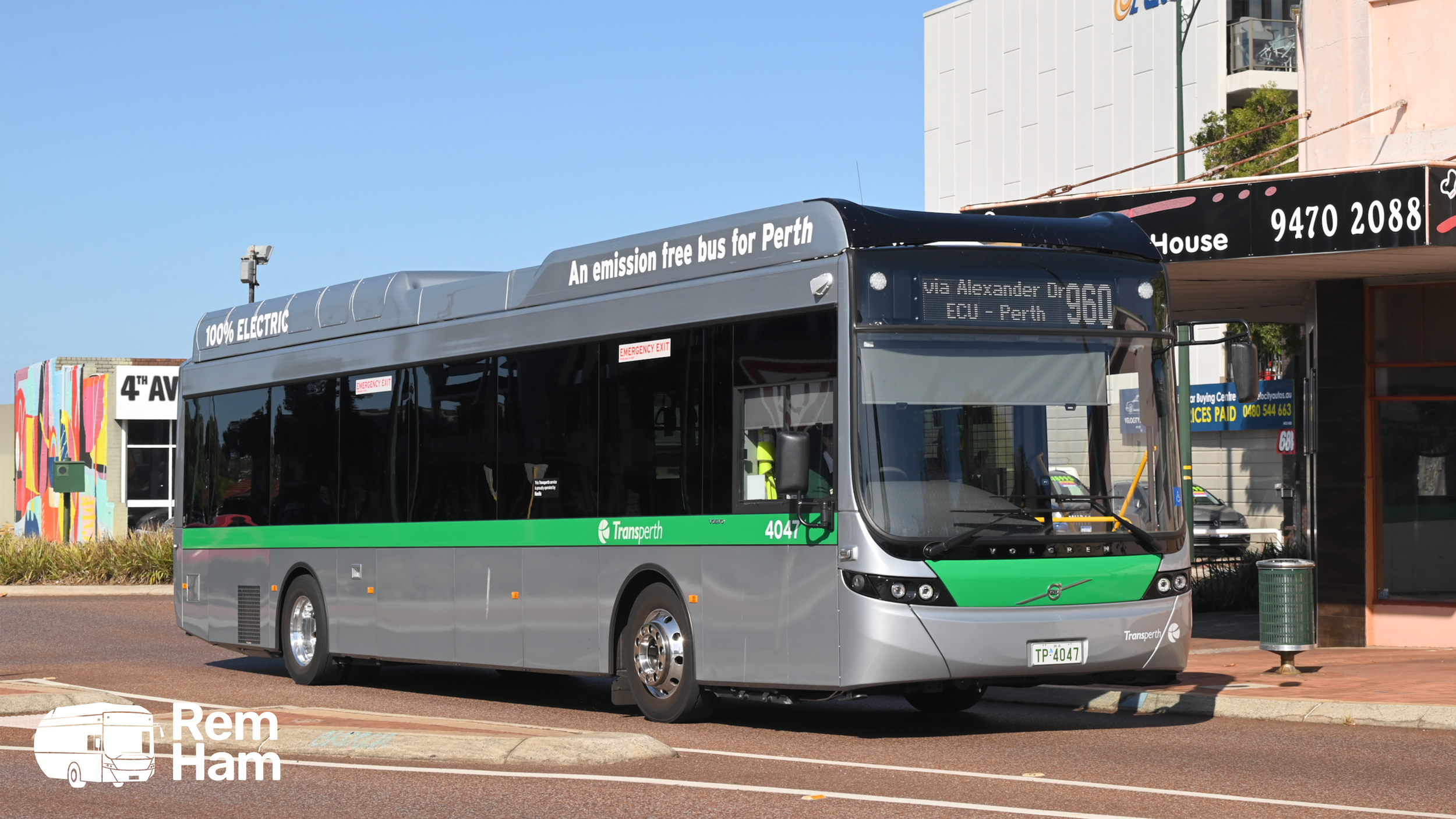 A modern electric bus with a green and silver color scheme, marked 'Transperth,' parked on the street near a building with a pink facade. The bus displays route information on a digital sign, and the words '100% Electric' and 'An emission free bus for Perth' are visible on the top. The bus is a Volvo model with a low-floor design.