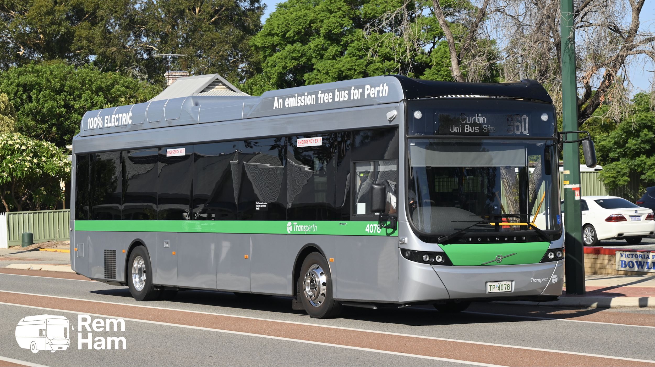 A modern electric bus with green and gray livery parked at a bus stop with trees and cars in the background. The bus displays route information for Perth, and has signs indicating it's 100% electric and emission free.