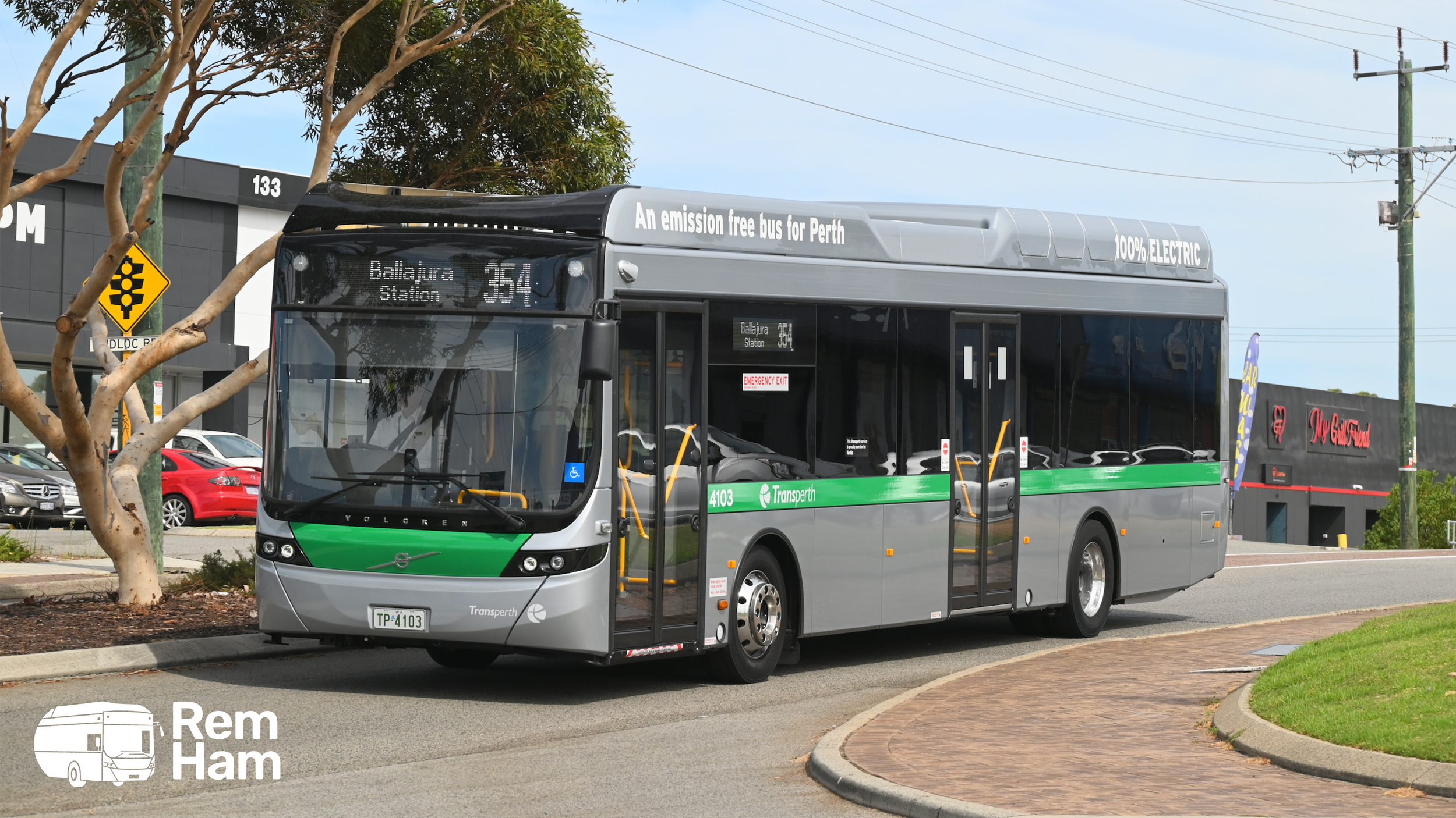 A modern electric bus with a green stripe, driven by Transperth, parked on a street near a sidewalk and trees, with buildings and parked cars in the background.