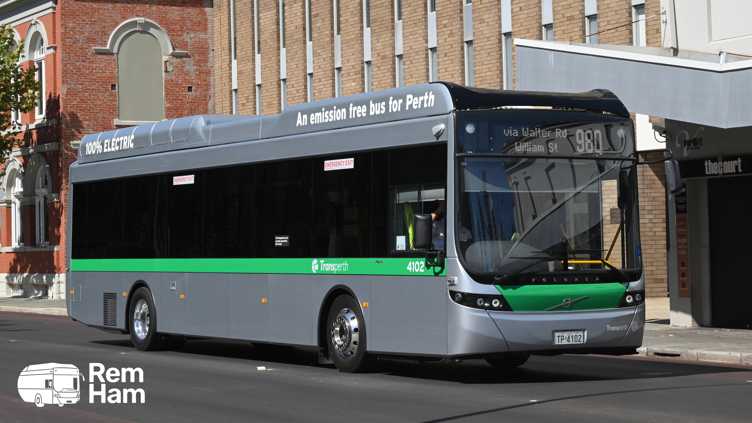A modern electric bus operated by Transperth, with a green and gray color scheme, driving on a city street in Perth, Australia, with buildings in the background. The bus displays the route number 980.