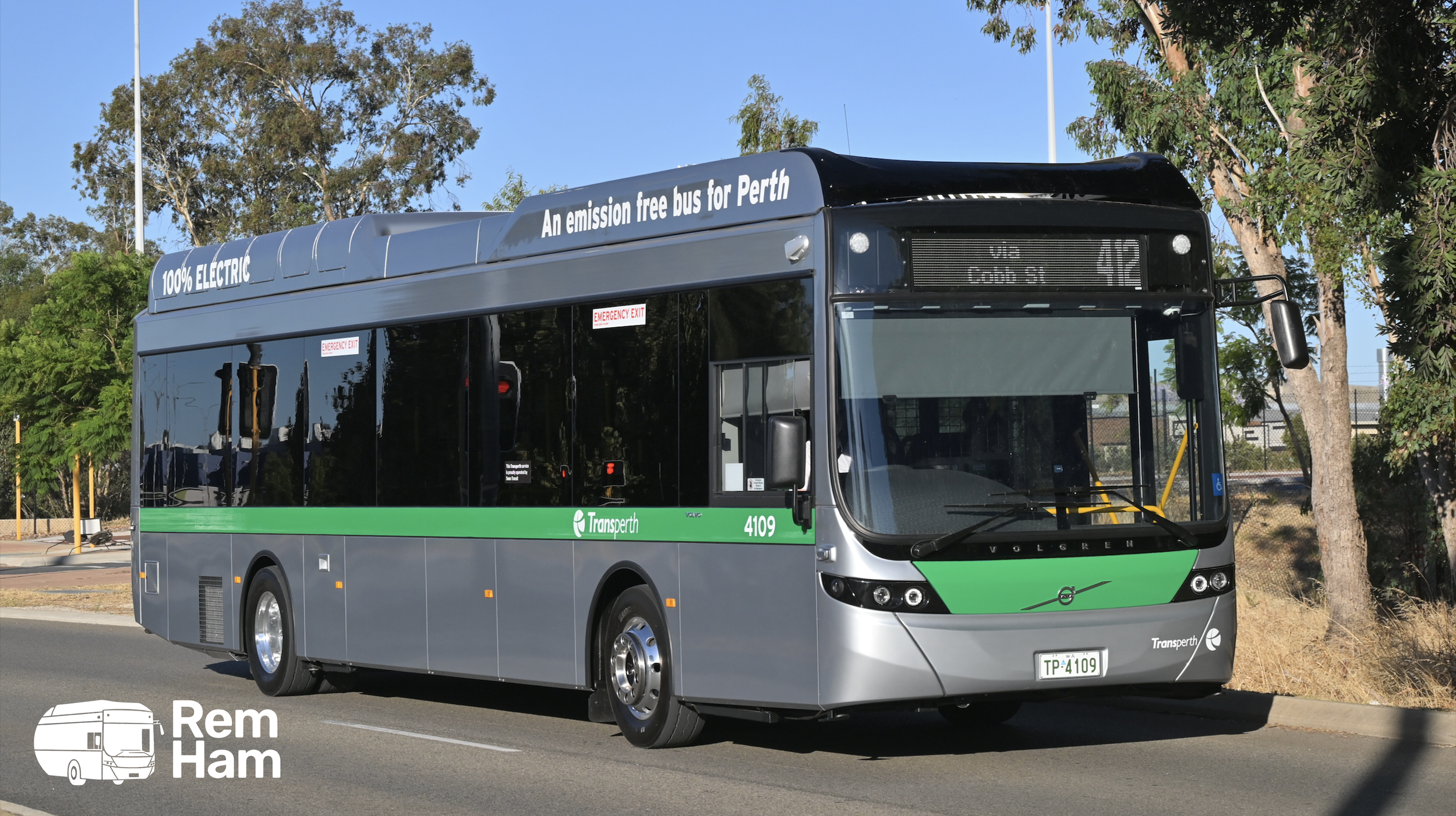A modern, gray electric bus with green accents, labeled as Transperth, parked on a street with trees and a blue sky in the background.