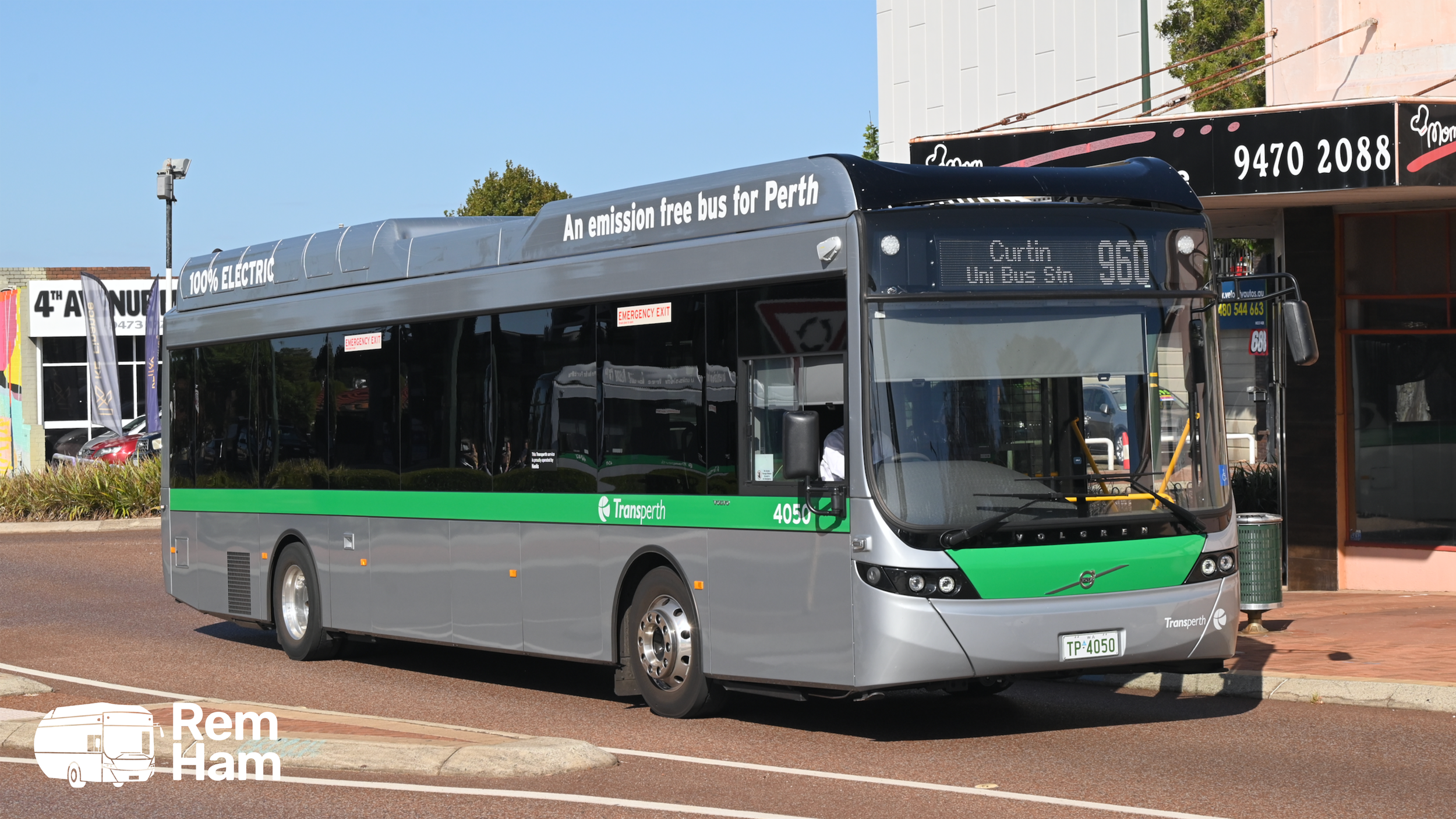 An electric bus with a silver and green color scheme parked at a bus stop on a city street, with signage indicating it is an emission-free bus for Perth.