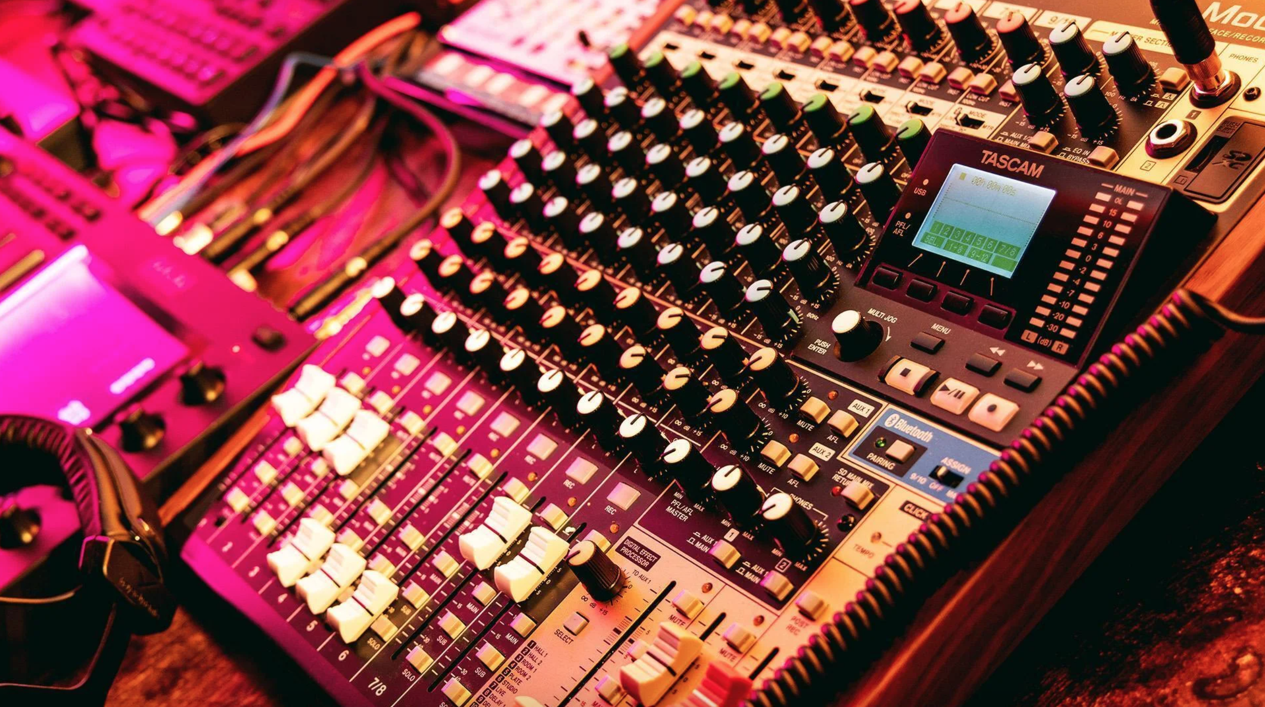 An audio mixing console with numerous knobs, sliders, and a small digital screen, illuminated with pink and warm lighting.