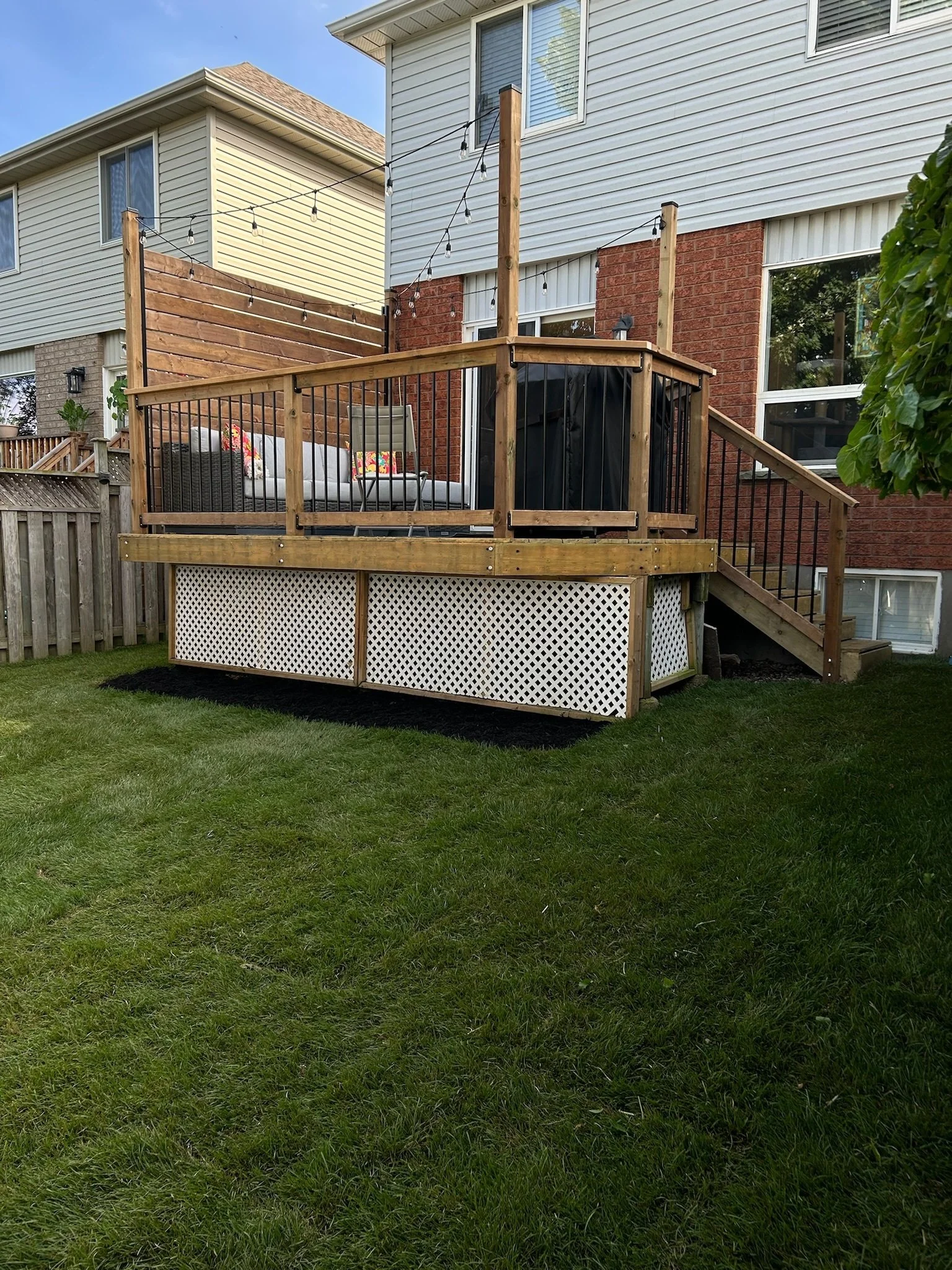Backyard wooden deck attached to a house, with outdoor furniture, string lights, and a railing, overlooking a grassy lawn.