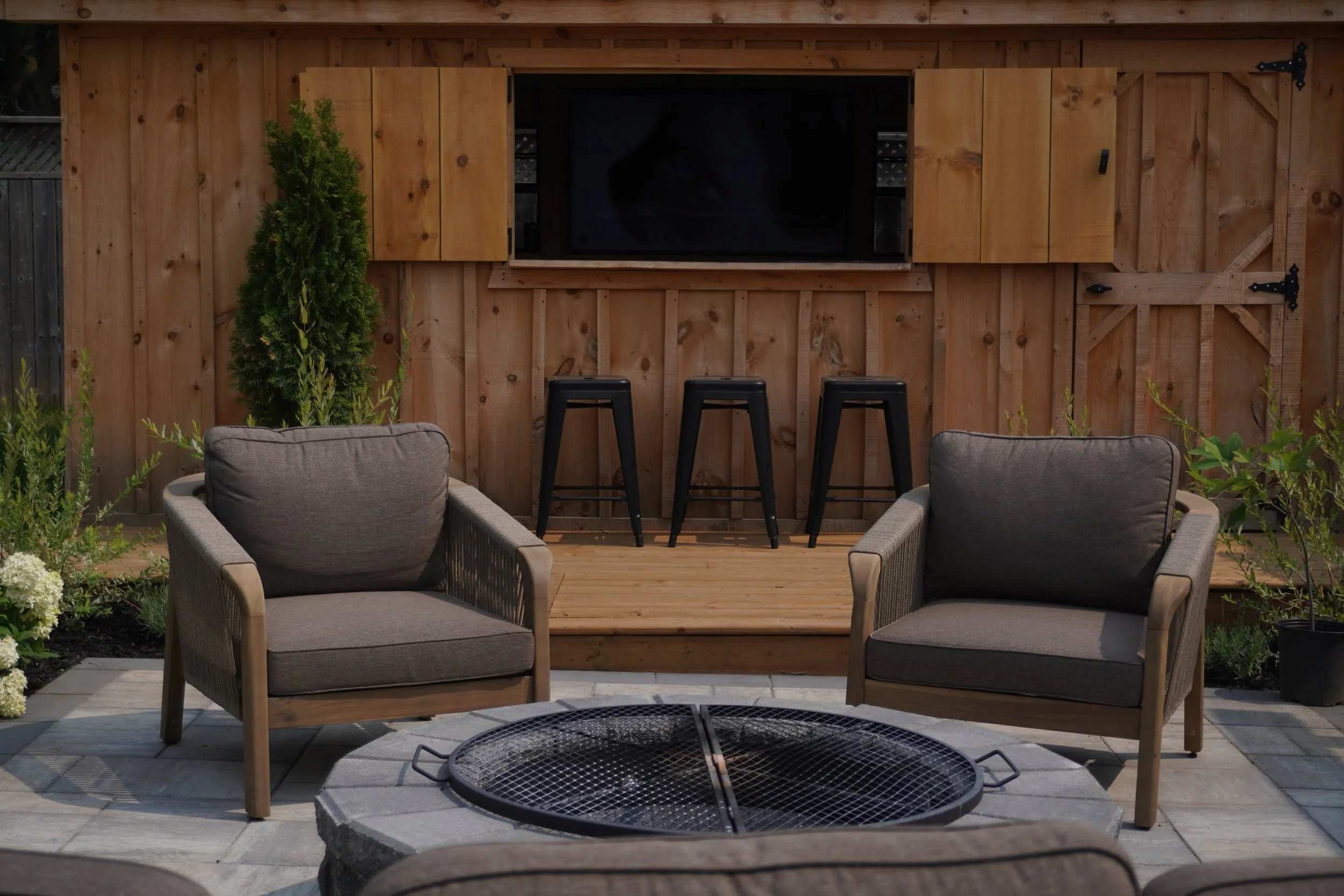 Backyard patio with gray cushioned chairs, a wood-fired fire pit, and a wooden shed with closed shutters and a window.