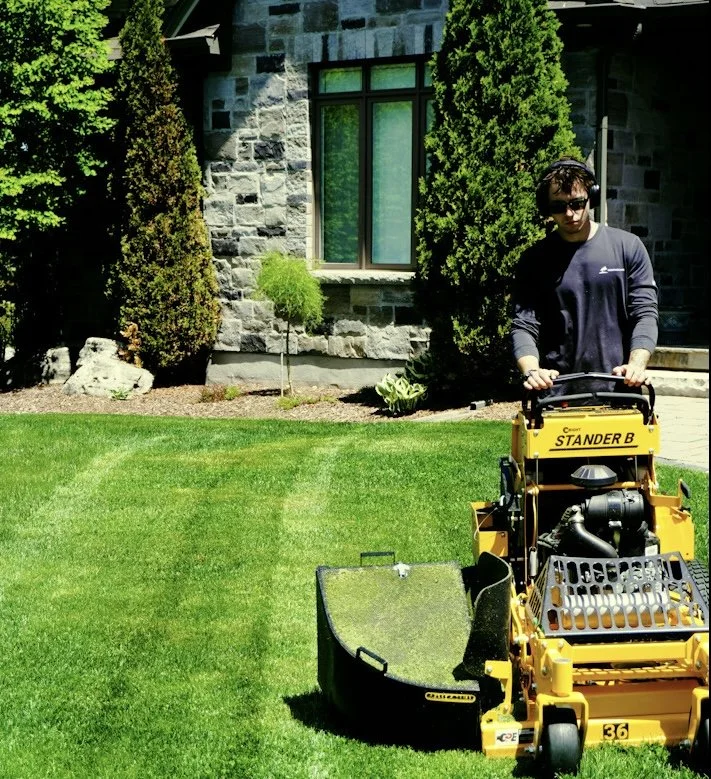 A man operating a yellow Stander B lawn mower on a well-maintained green lawn in front of a stone house with a large window and tall trees.