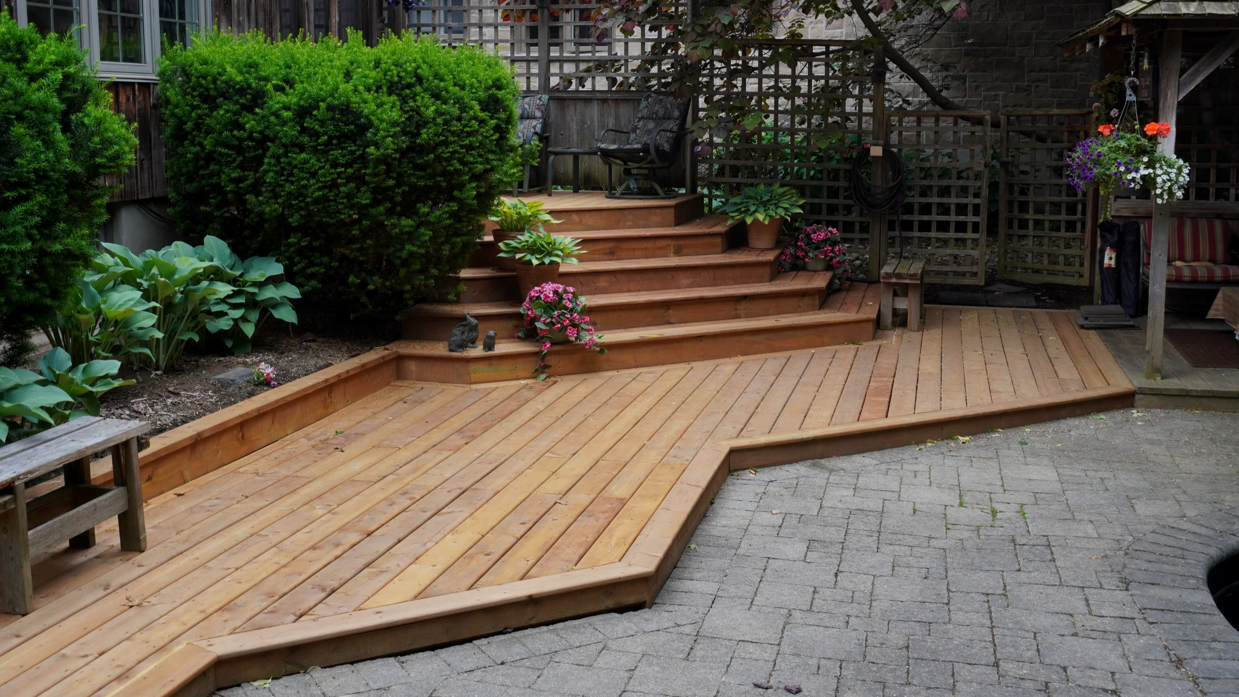 A backyard patio with a wooden deck, stairs, and garden with green shrubs, flowering plants, and outdoor furniture.