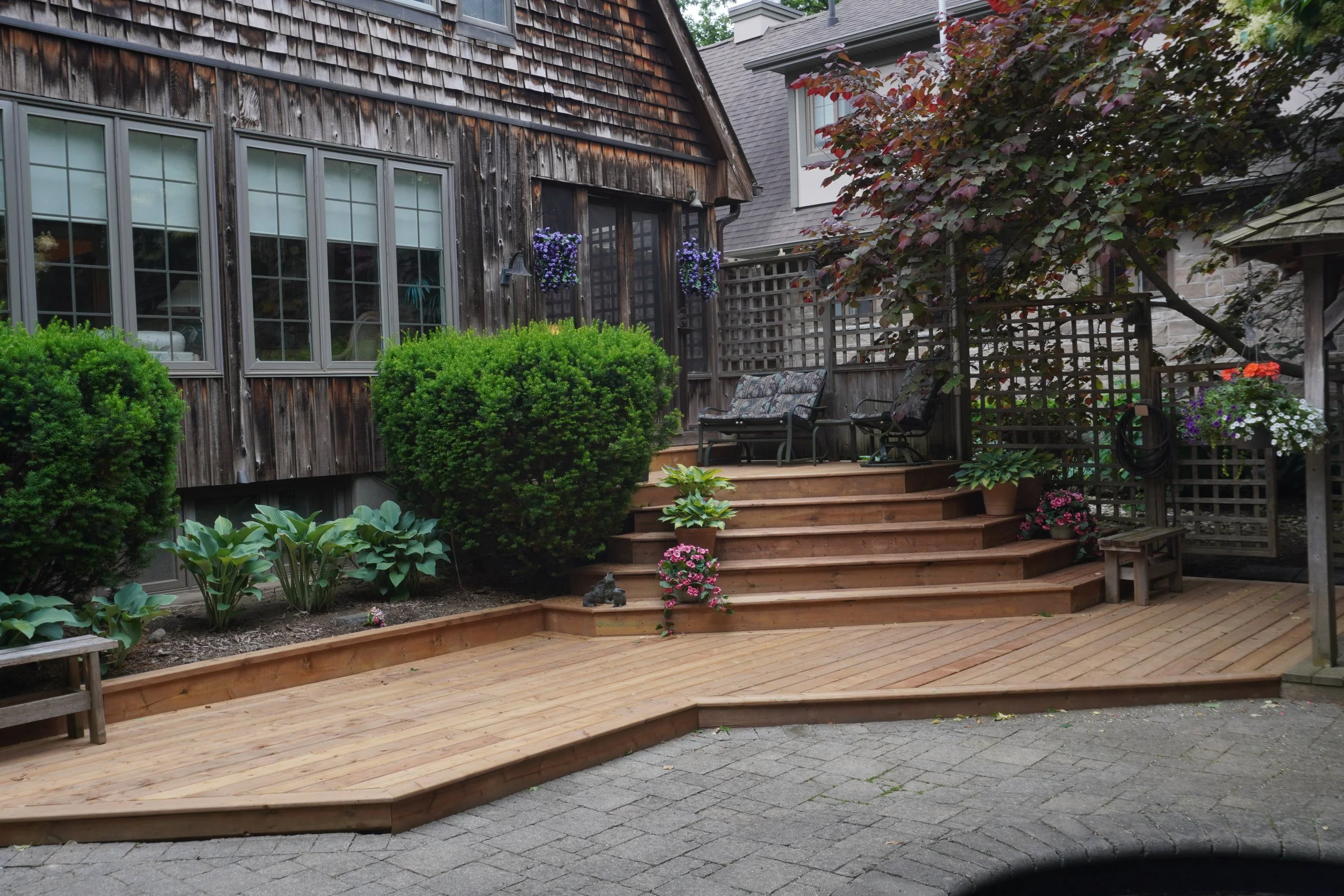 A backyard patio with a wooden deck, steps, and green shrubbery, featuring chairs, potted flowers, and climbing plants.