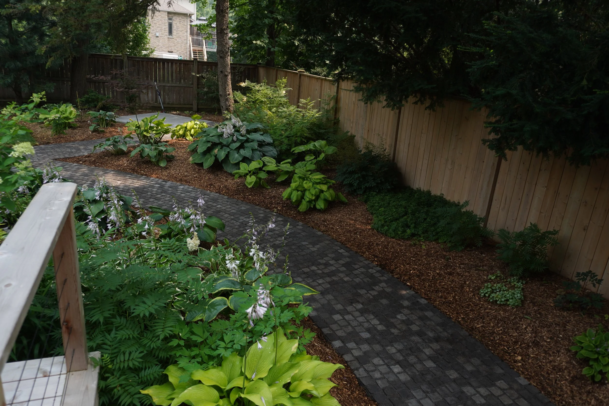 A landscaped backyard with a cobblestone pathway, various green plants, and trees, bordered by a wooden fence. A house with a stone exterior and a staircase leading up to a deck are visible in the background.