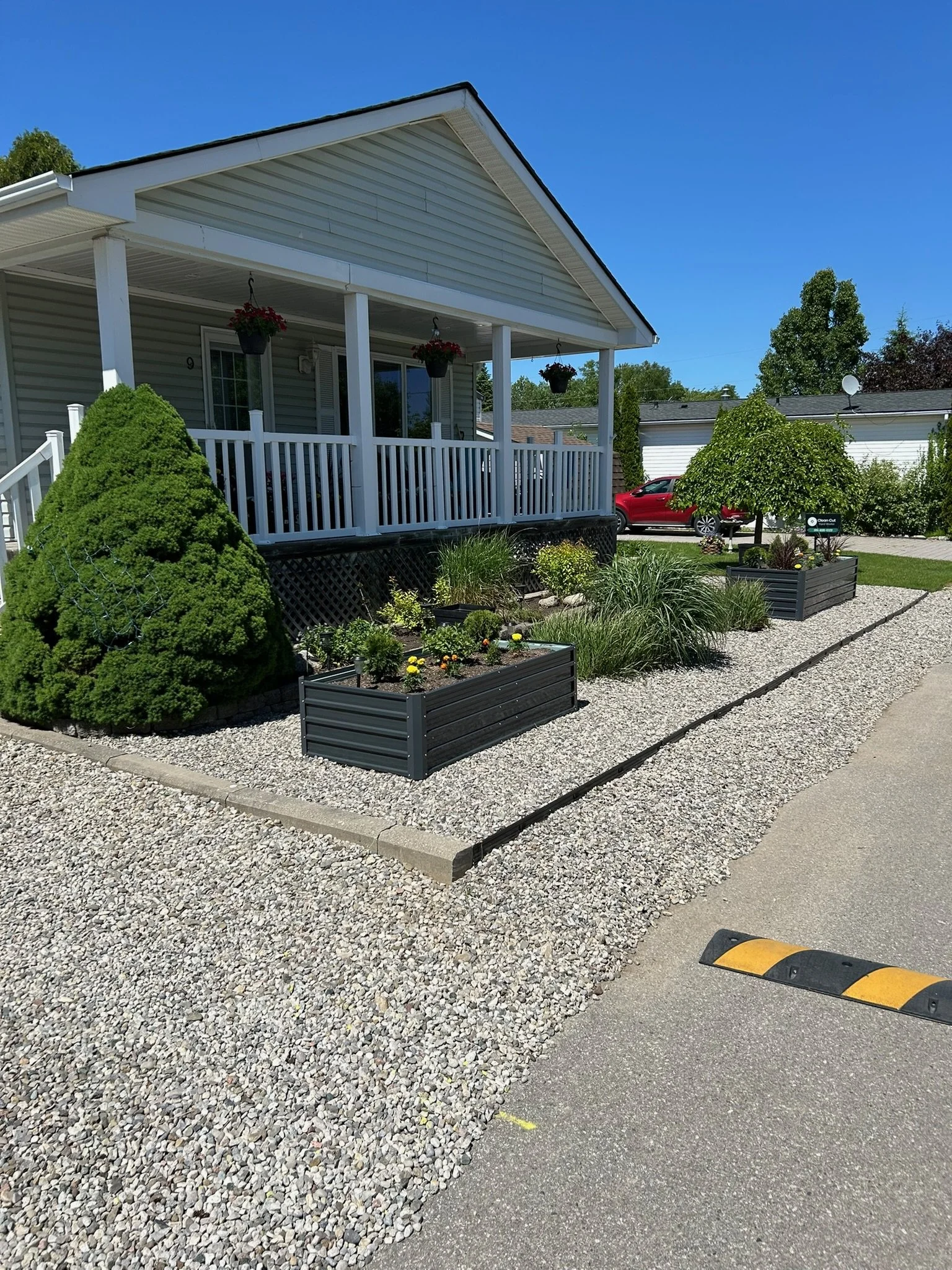 A front yard with a house featuring a porch, hanging flower baskets, and landscaped garden beds with shrubs and flowers, bordered by gravel, with a parking lot in the foreground and a bright blue sky overhead.