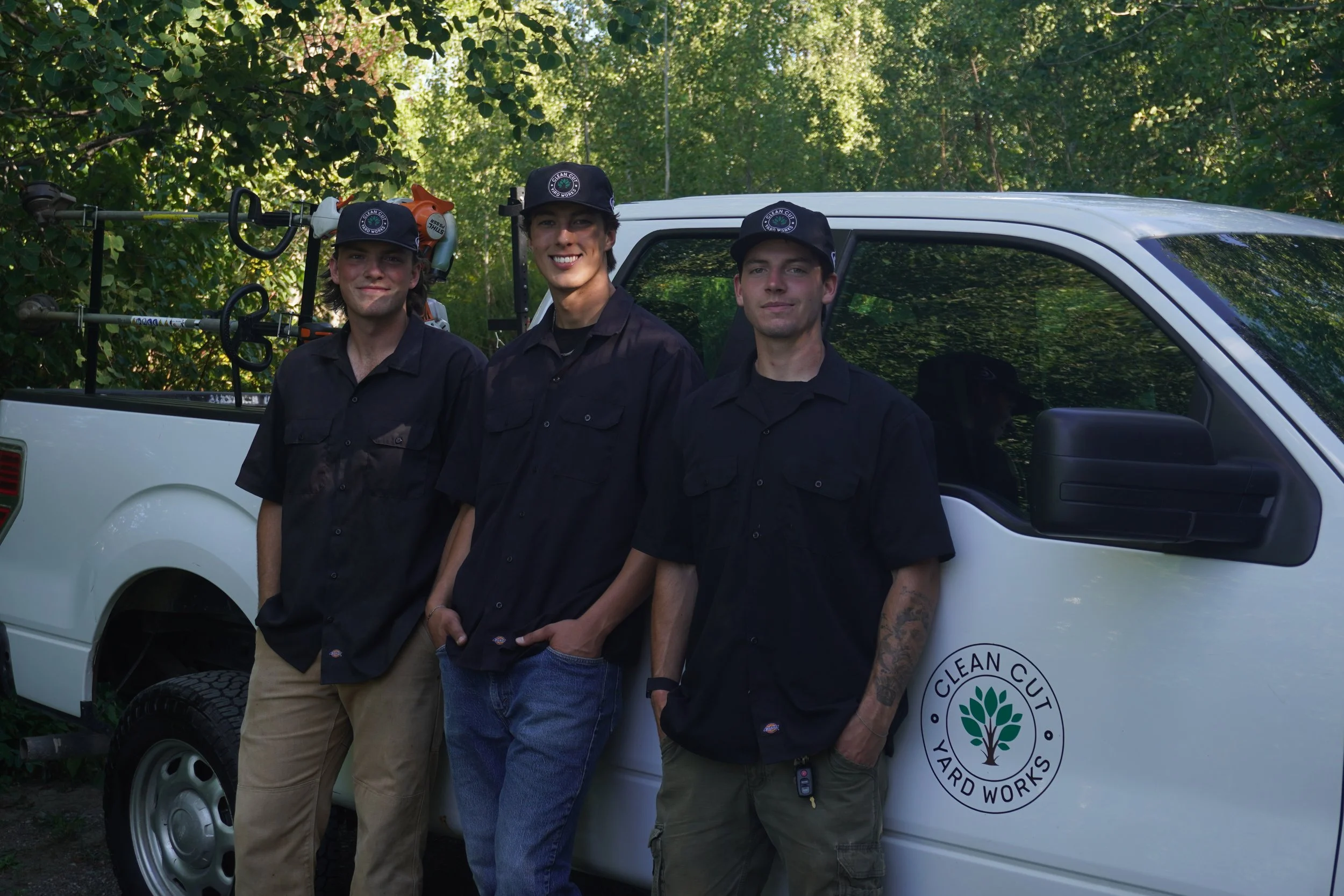 Three young men in black work shirts and caps standing in front of a white pickup truck labeled 'Clean Cut Yard Works'. They are outdoors with lush green trees in the background, and tools are visible in the truck bed.