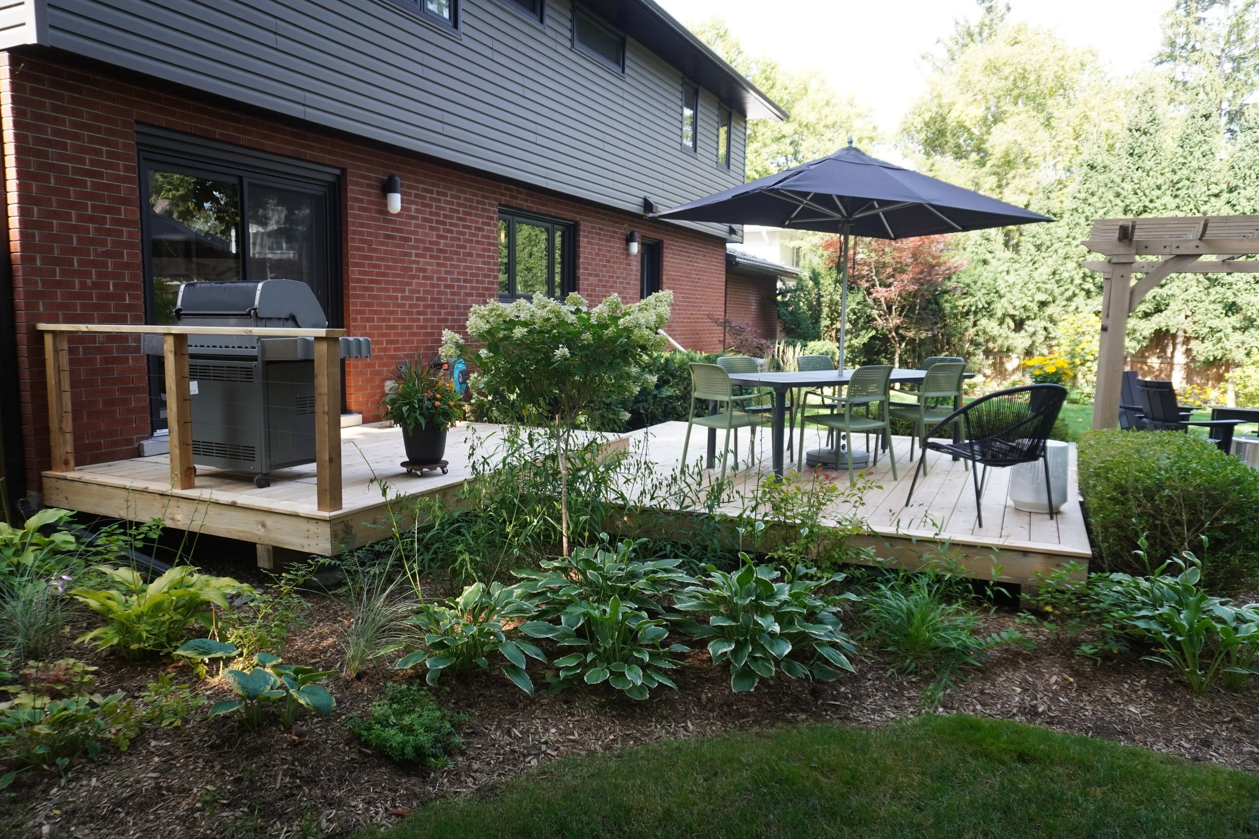 Backyard patio with outdoor dining set under a large umbrella, surrounded by garden plants and trees, adjacent to a brick and siding house.