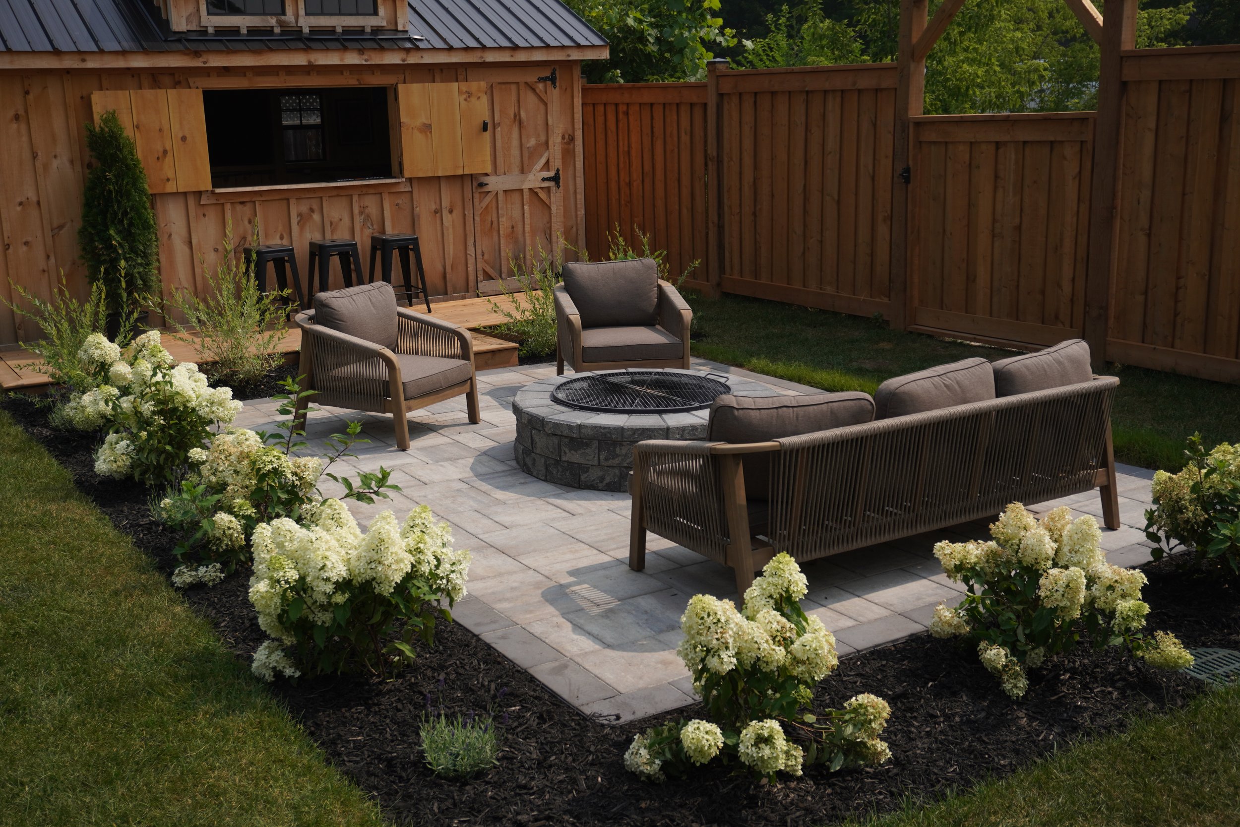 A backyard patio with outdoor seating, a fire pit, garden plants and flowers, and a wooden privacy fence with a small storage shed.