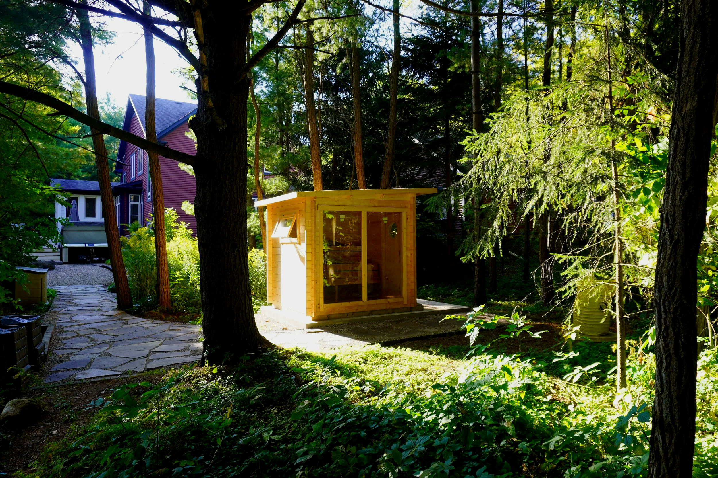 A small wooden structure with glass doors, situated in a wooded backyard surrounded by trees, plants, and a stone pathway.