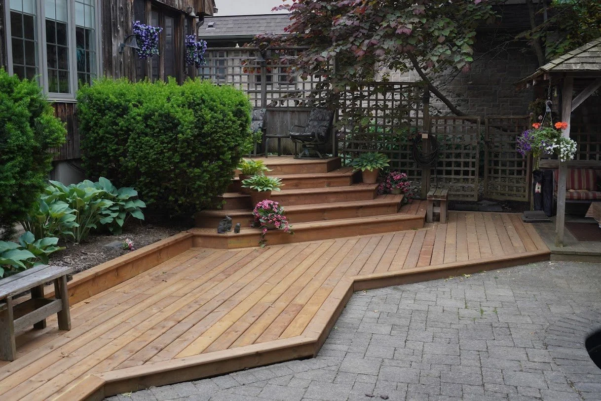 Backyard with a wooden deck, steps, green shrubs, potted plants, and outdoor furniture.