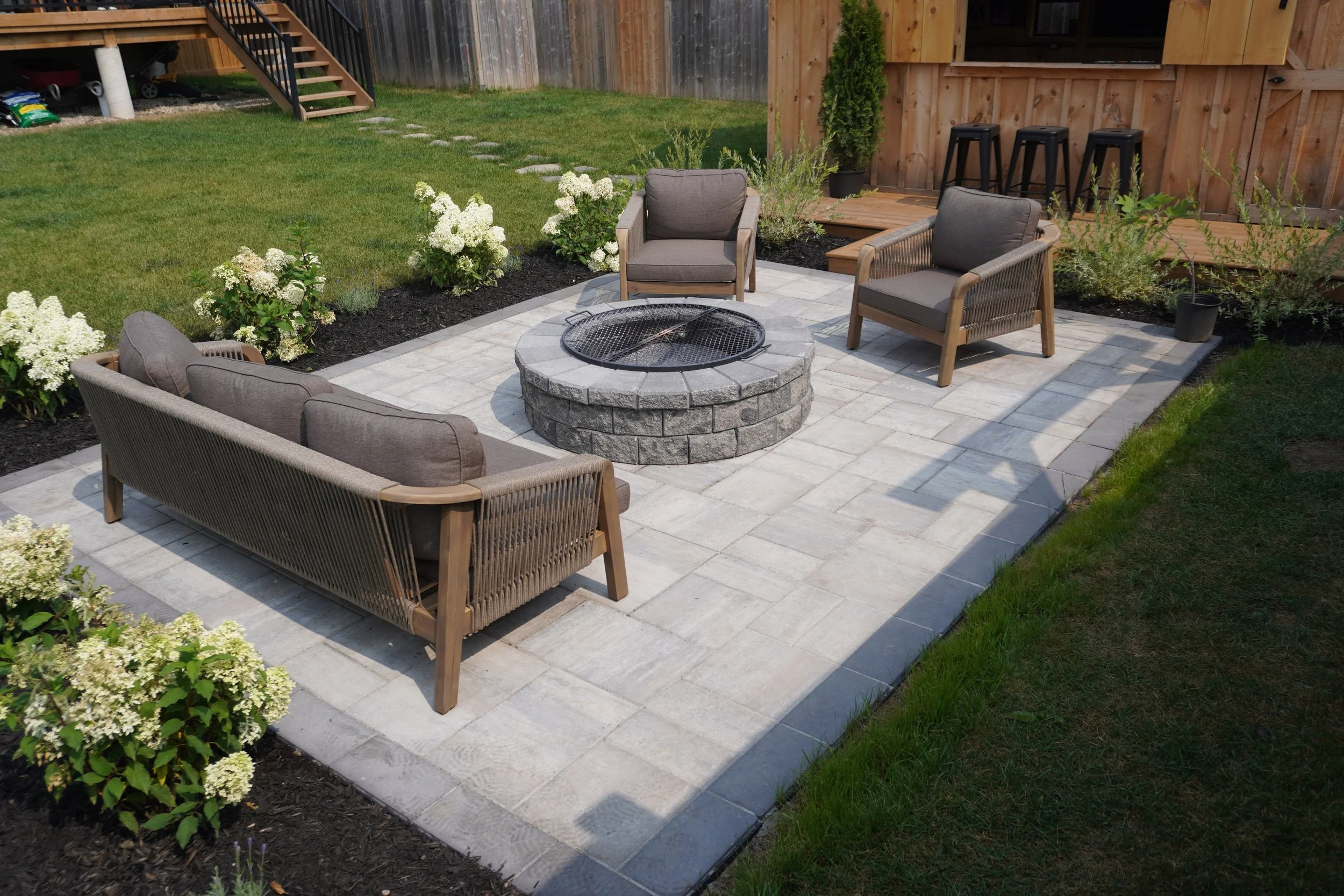 A backyard patio with three gray cushioned chairs around a circular stone fire pit. The patio is paved with light-colored stone tiles and surrounded by a grassy lawn with white flowering bushes. In the background, a wooden fence, a raised wooden deck