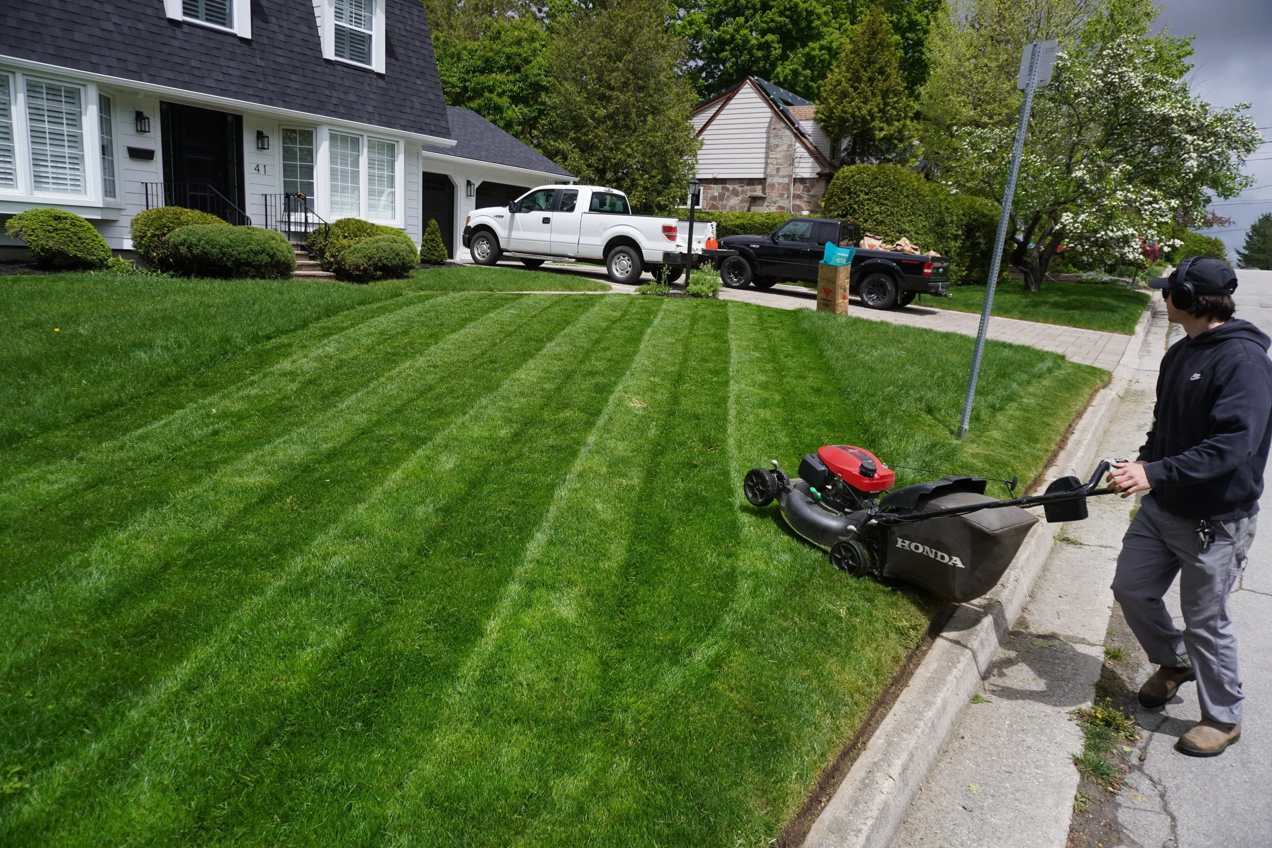 A person operating a lawn mower on a residential front lawn with freshly cut grass; houses, trees, and parked trucks are in the background.