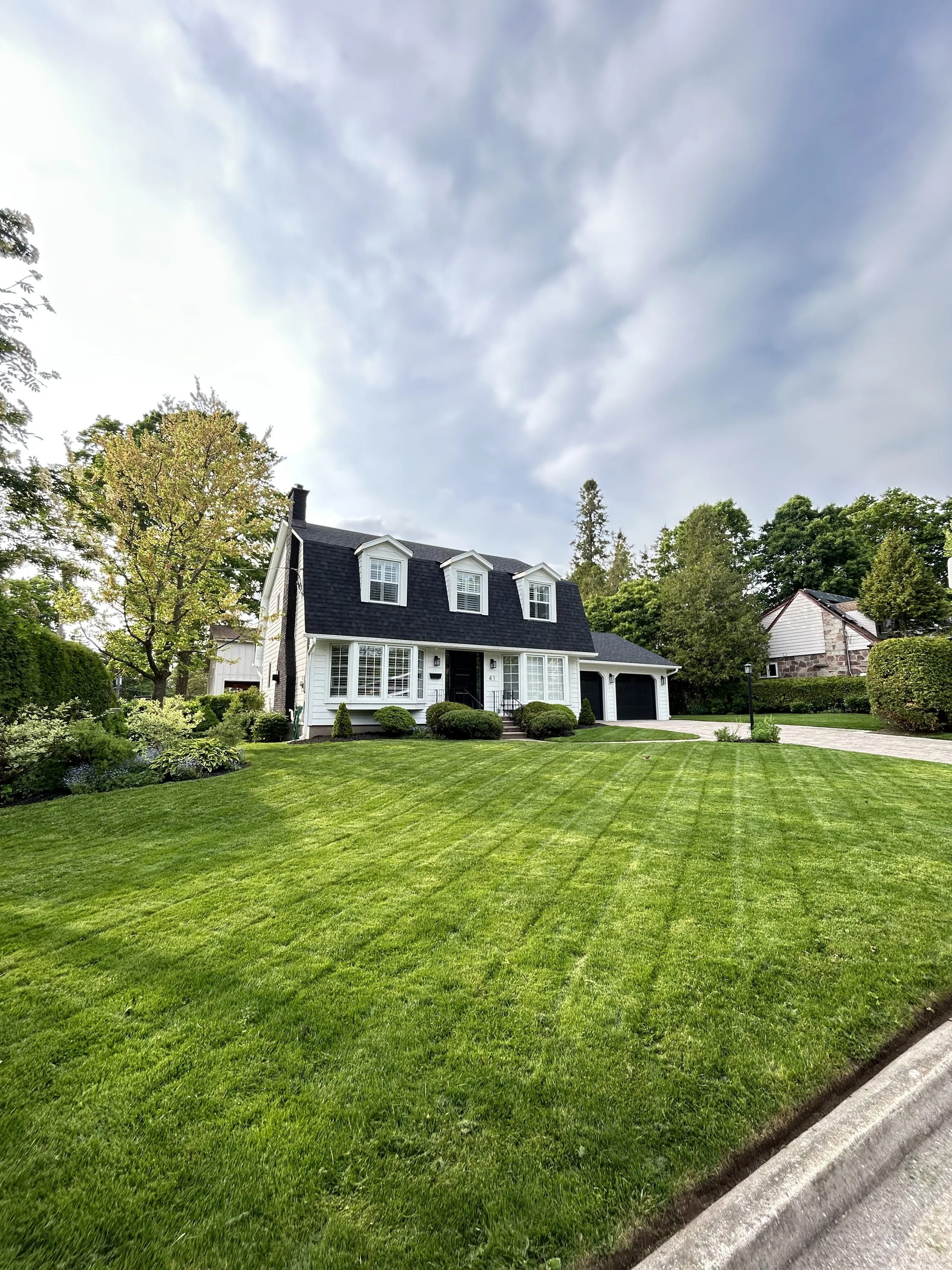 A white house with black roof windows and a courtyard, surrounded by well-maintained green lawn and trees, under a partly cloudy sky.