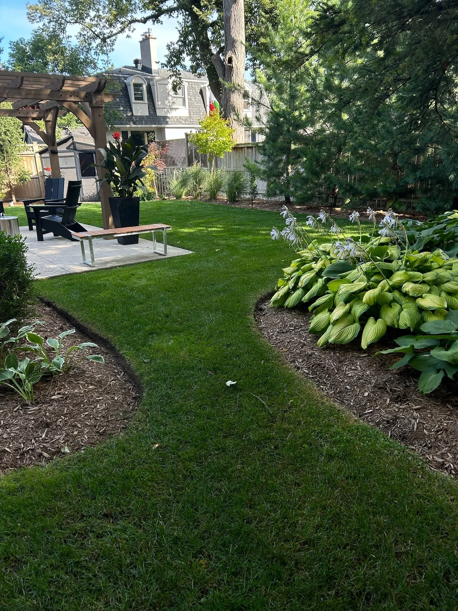 A well-maintained backyard garden with a grassy lawn, flower beds, and a seating area with chairs and a bench. There is a large tree, shrubs, and neighboring houses in the background.