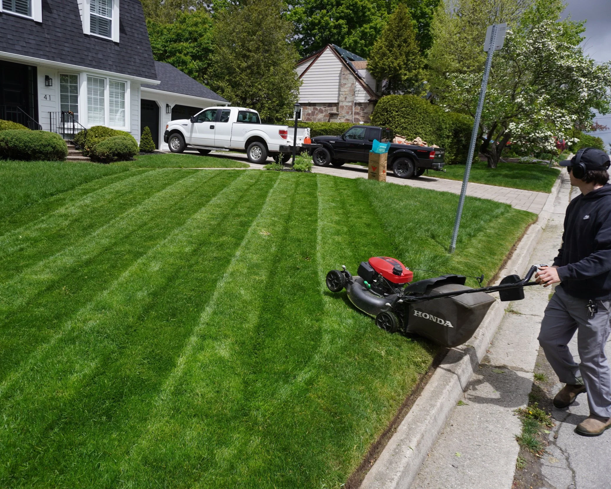 Person with dark hair wearing a cap operating a lawn mower on a grass lawn next to a sidewalk and house with two trucks parked in the driveway and trees in the background.