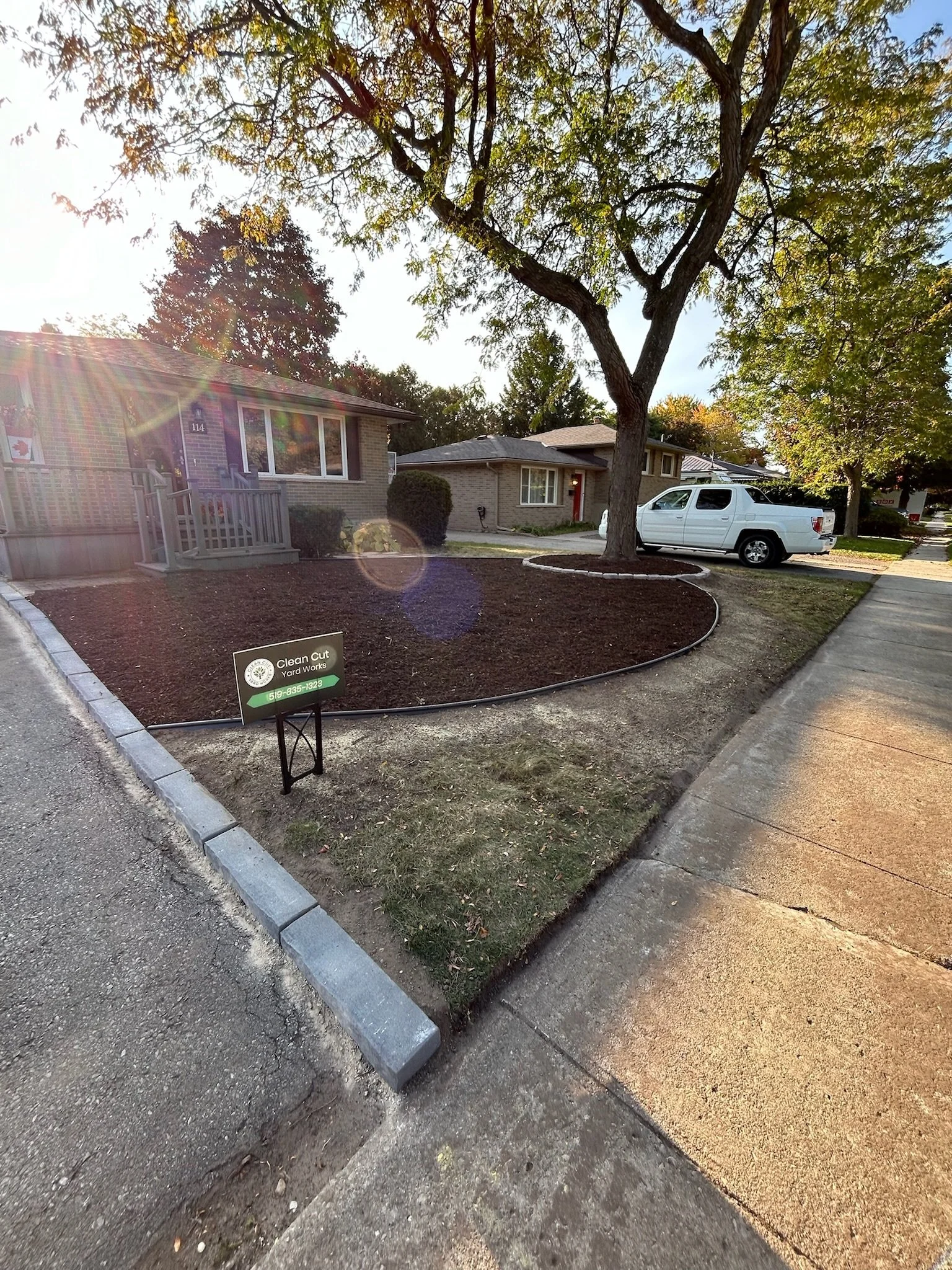 Front yard with freshly mulched flowerbed, large tree, sidewalk, and a white pickup truck parked on the street in a residential neighborhood.