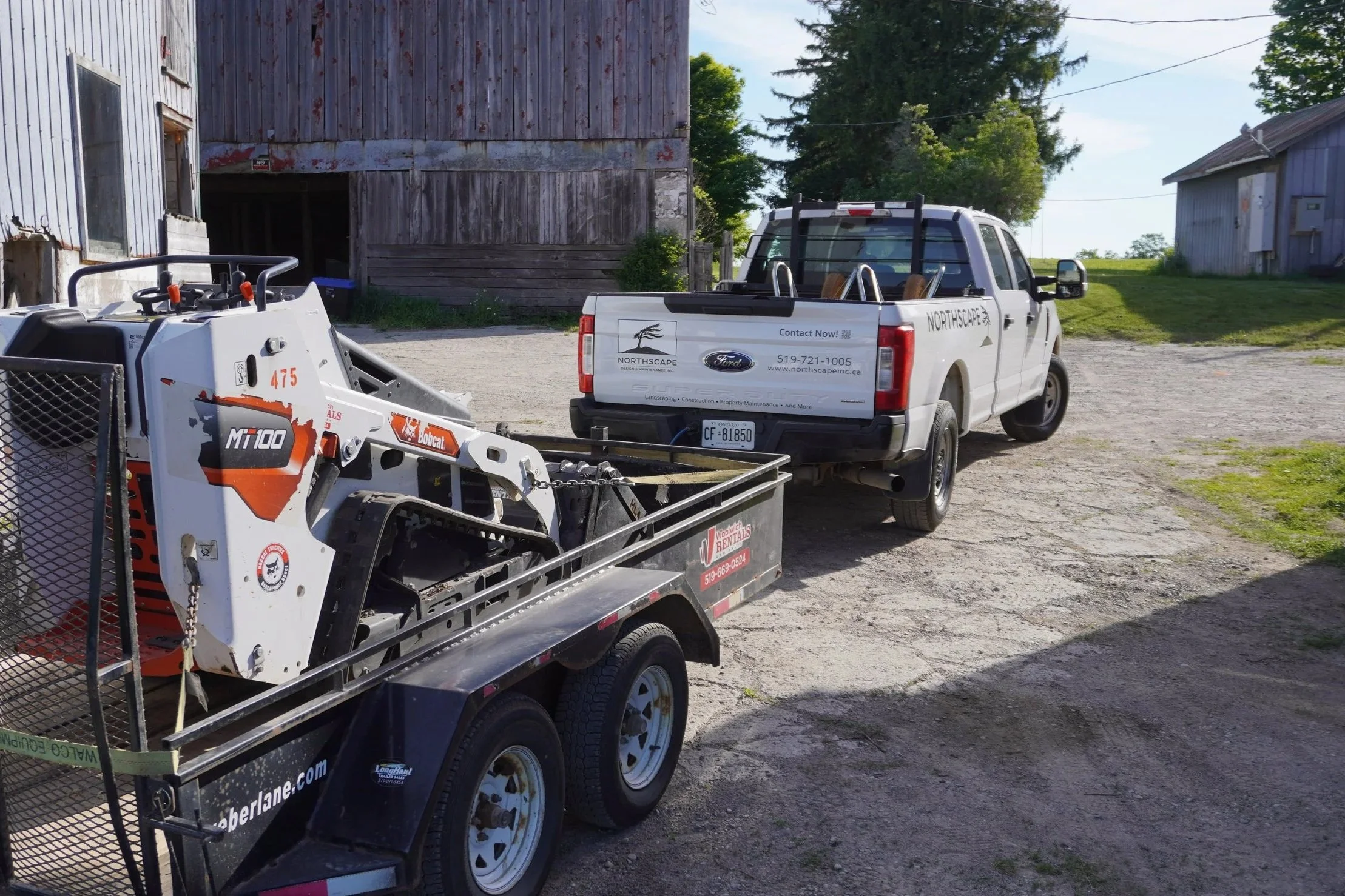 A white pickup truck from Northscape, parked next to a trailer with equipment, in front of an old wooden barn on a gravel lot with green trees and a blue sky.