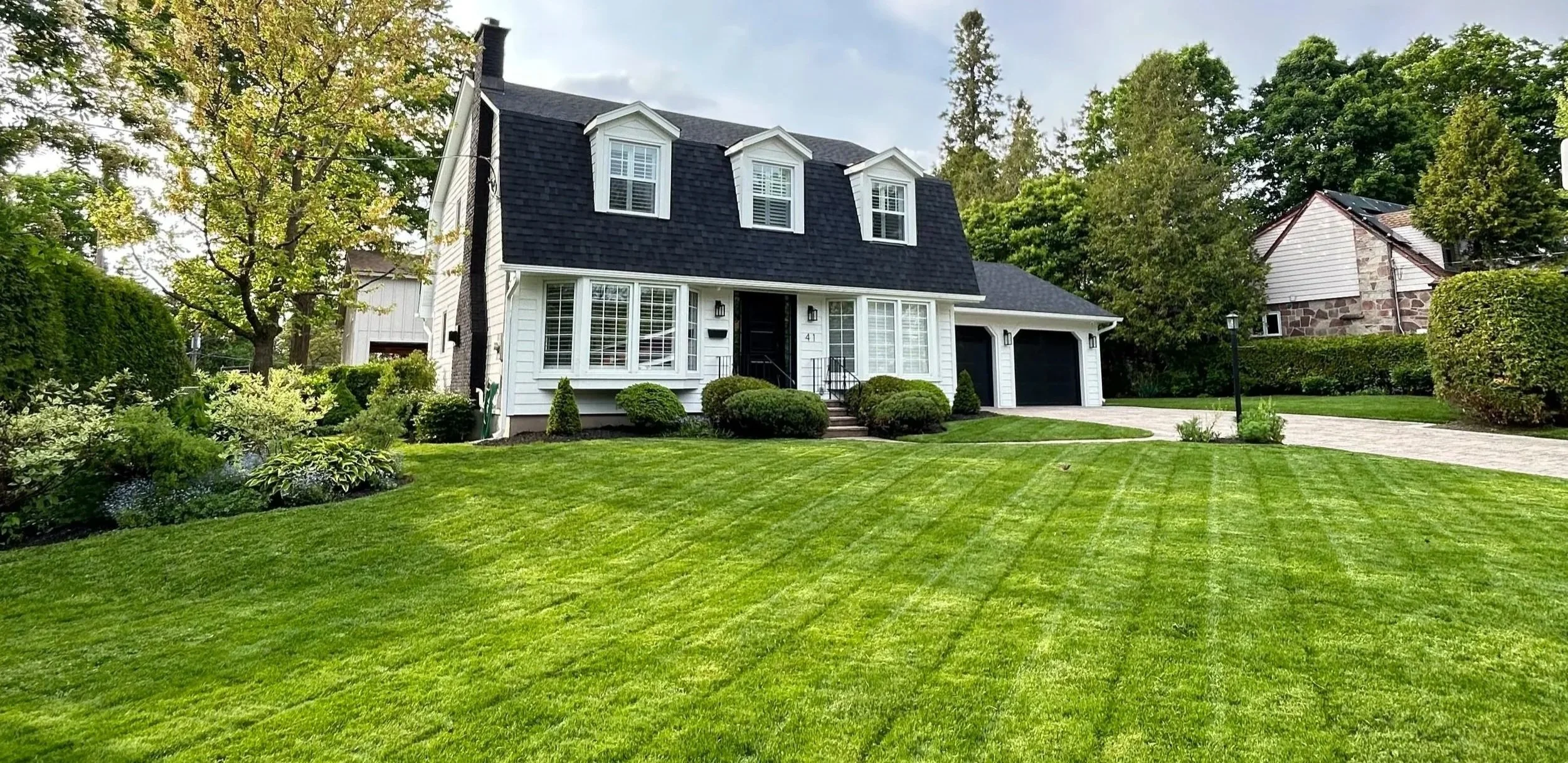 A white two-story house with black roof and three dormer windows, surrounded by green trees and lawn, with a paved driveway on the right side and outdoor lighting fixtures.