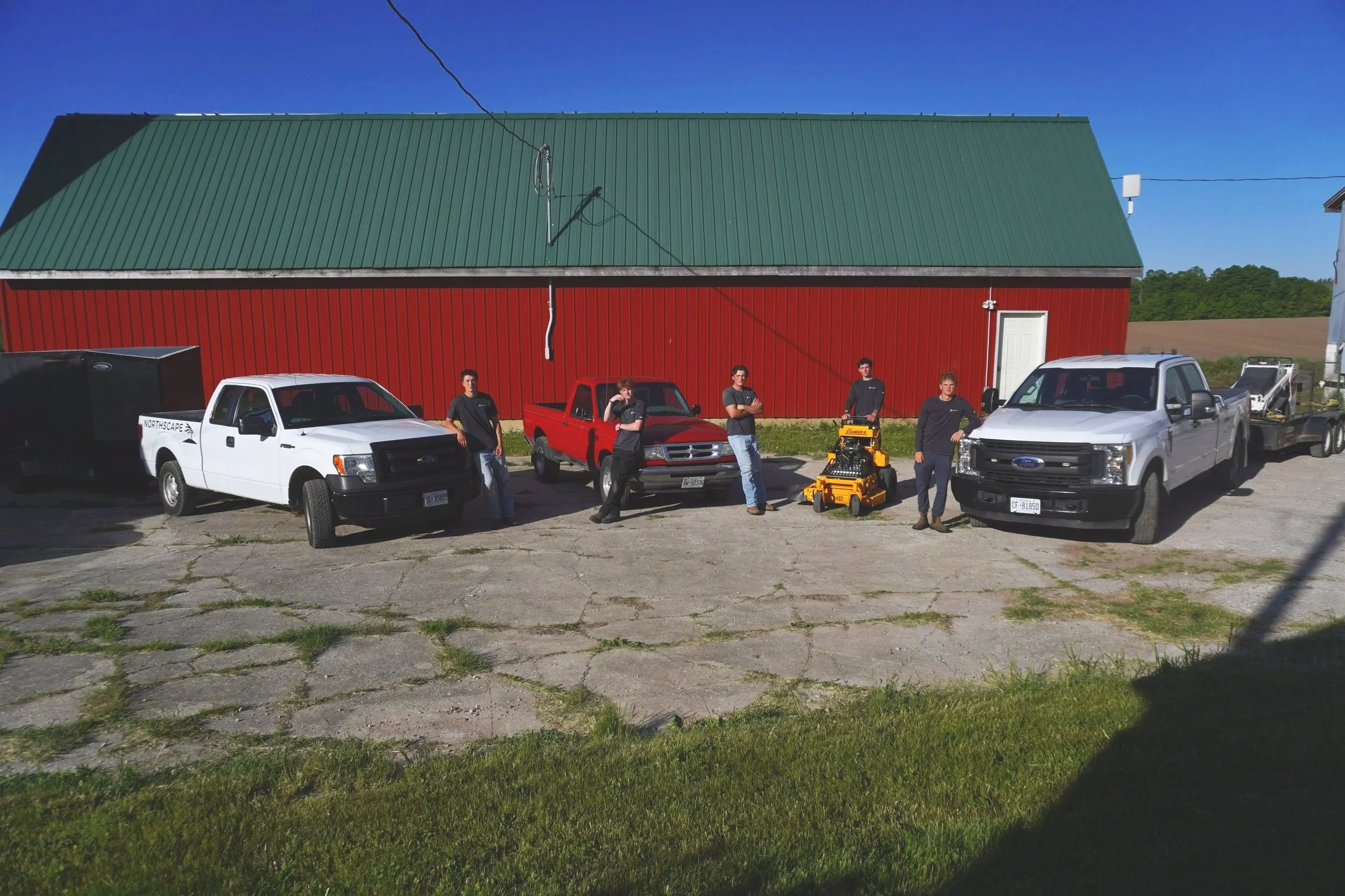 Six young men standing in front of a large red barn with a green metal roof, with vehicles parked nearby and a clear blue sky overhead.