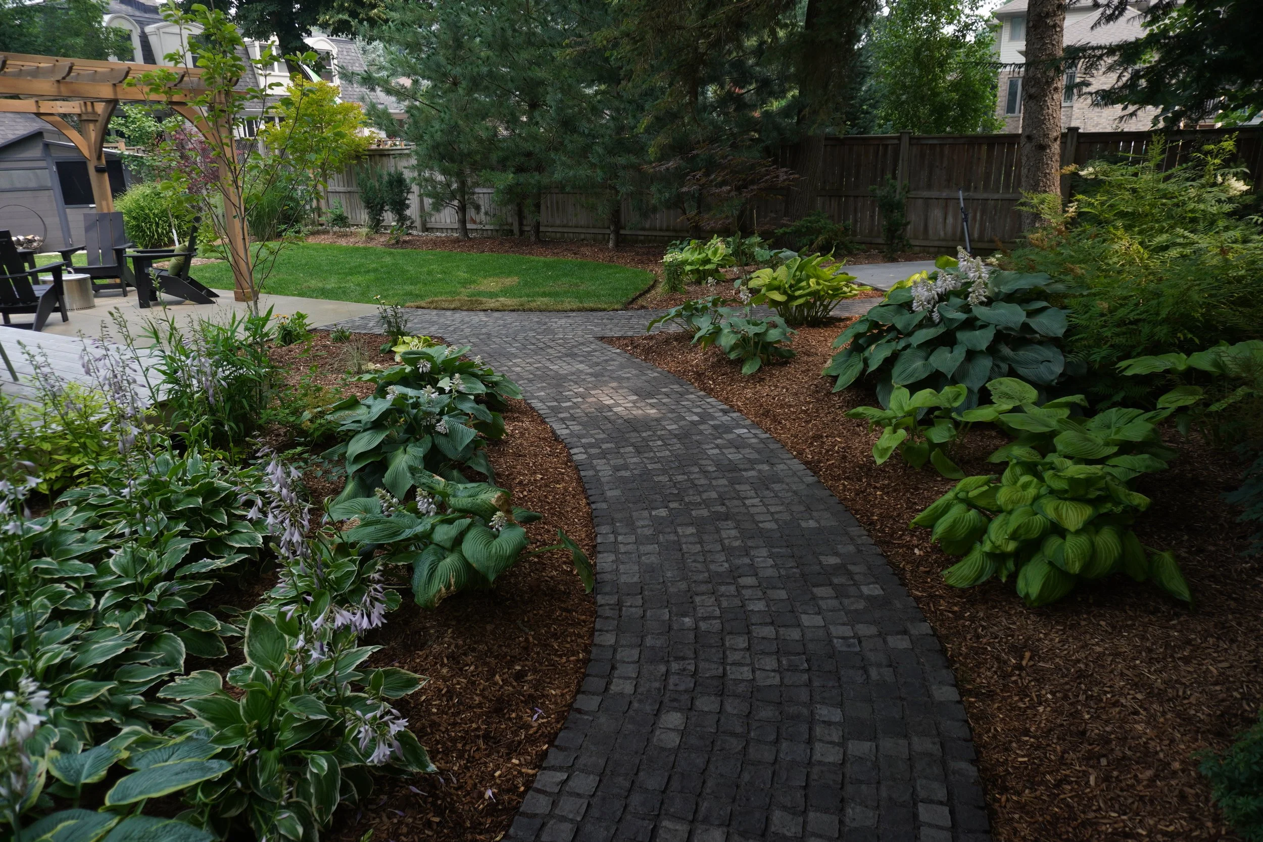 A backyard garden with a curved stone pathway, surrounded by lush green plants, trees, and a wooden fence.