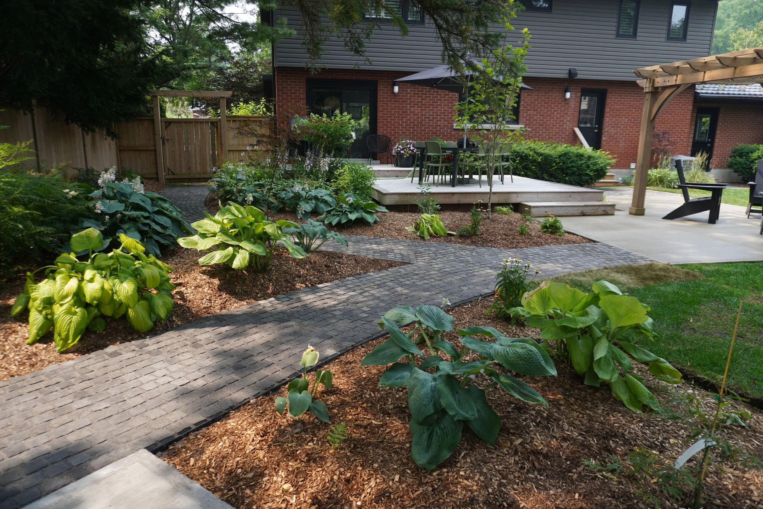 Residential backyard with a wooden deck, outdoor dining table with chairs, a black chair, a pergola, lush green plants, and a house with brick and gray siding.