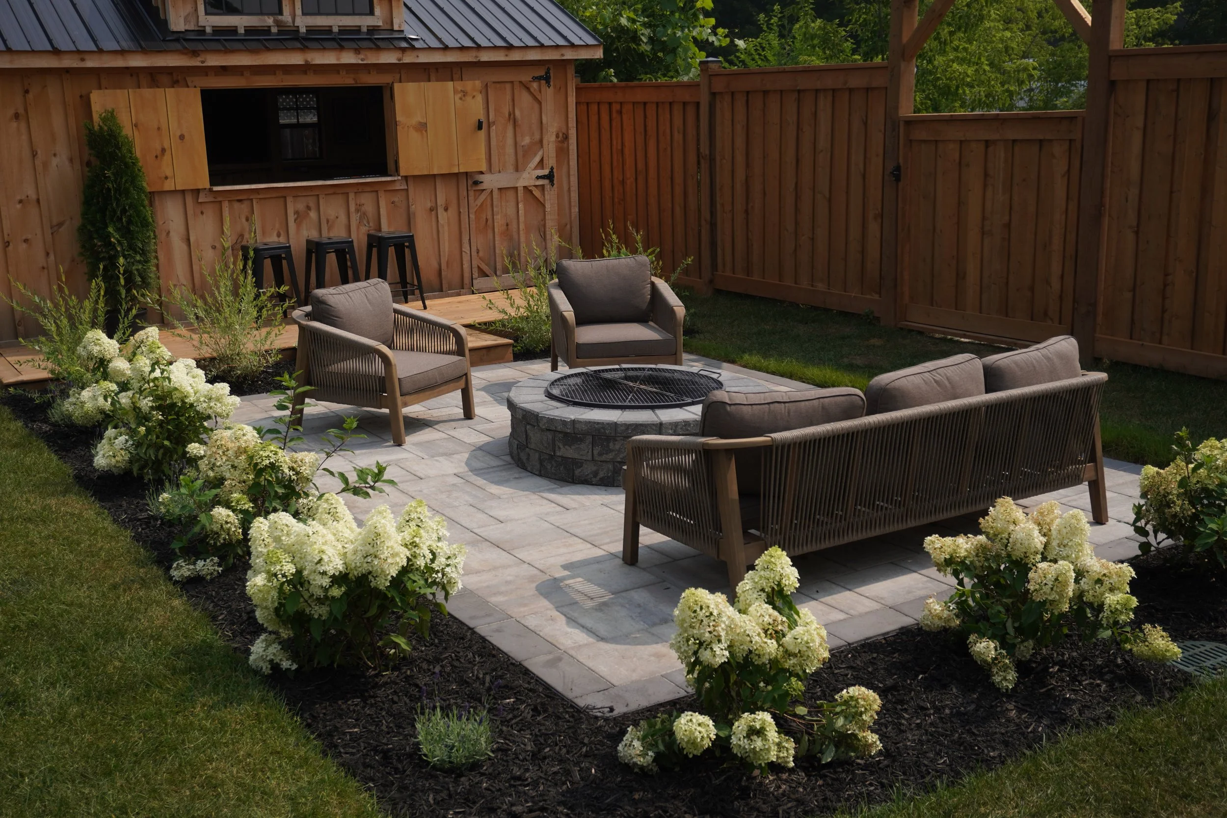 A backyard patio with outdoor furniture, a fire pit, flowering bushes, and a wooden privacy fence.