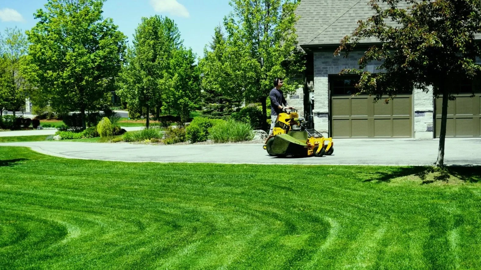 A person using a yellow lawn mower to cut grass in a green backyard with trees and a house in the background.