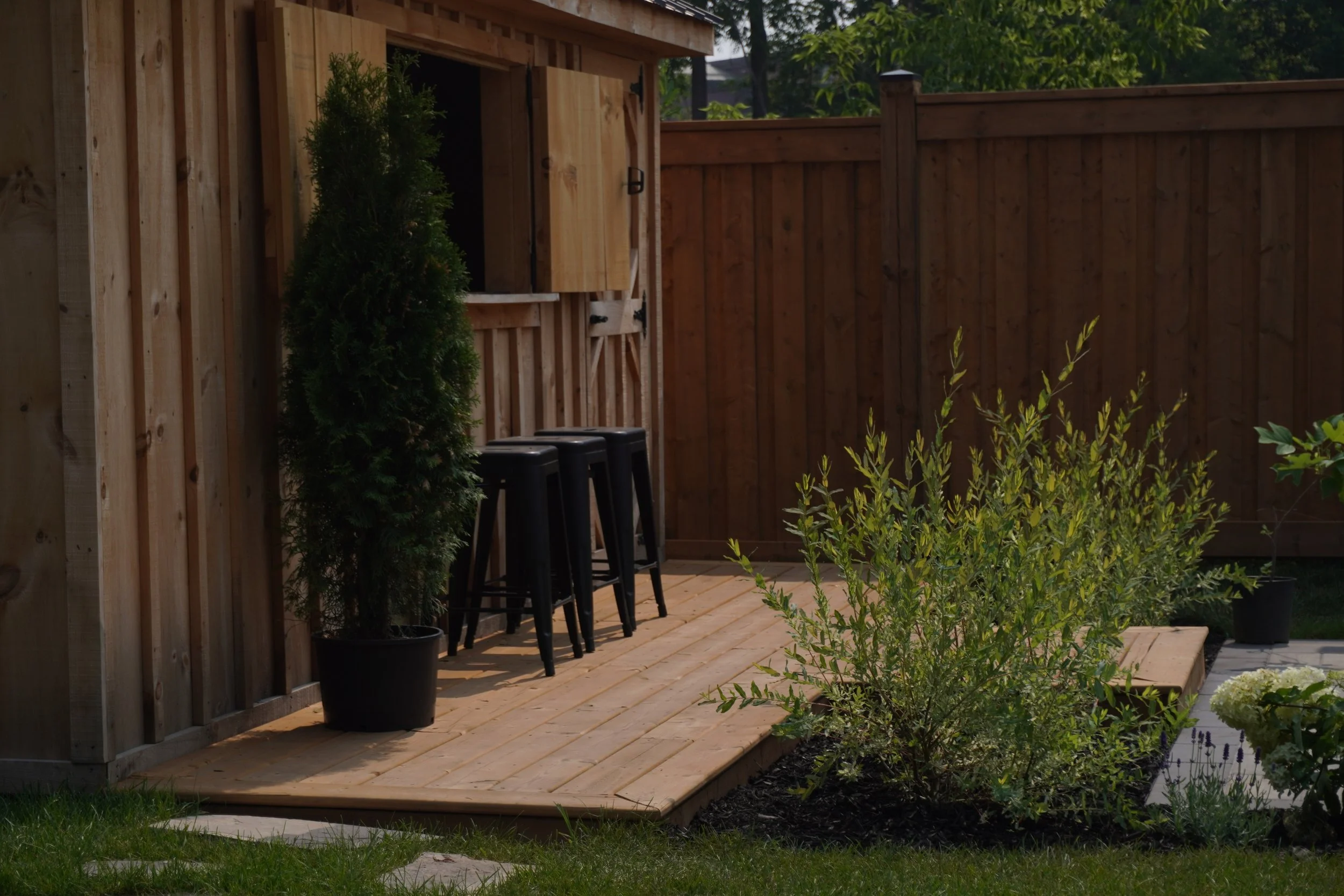 A backyard with a wooden privacy fence, a small wooden deck with three black stools, potted plants including a small evergreen, and various green bushes and trees.