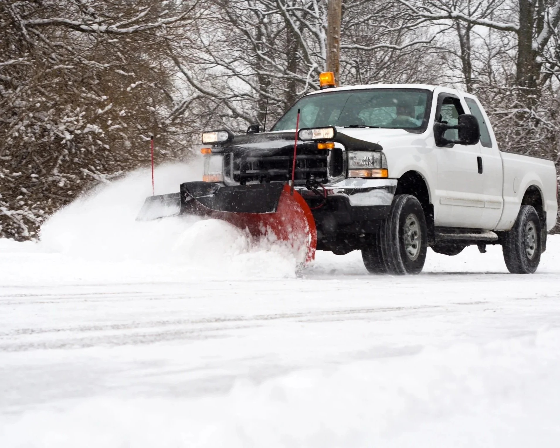 A white pickup truck equipped with a snow plow clearing snow from a road, with snow-covered trees in the background.