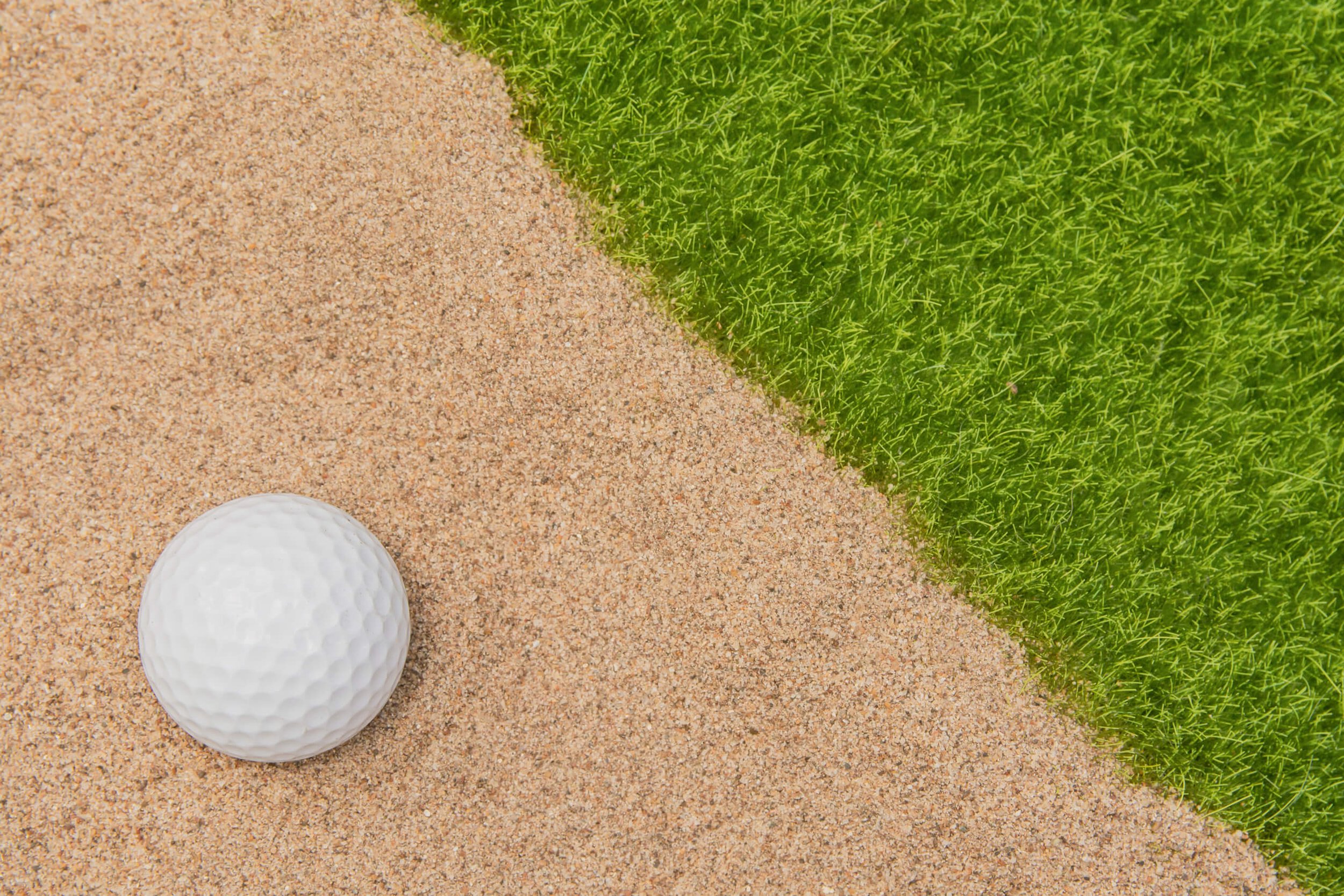 A golf ball on sand next to a patch of green grass.
