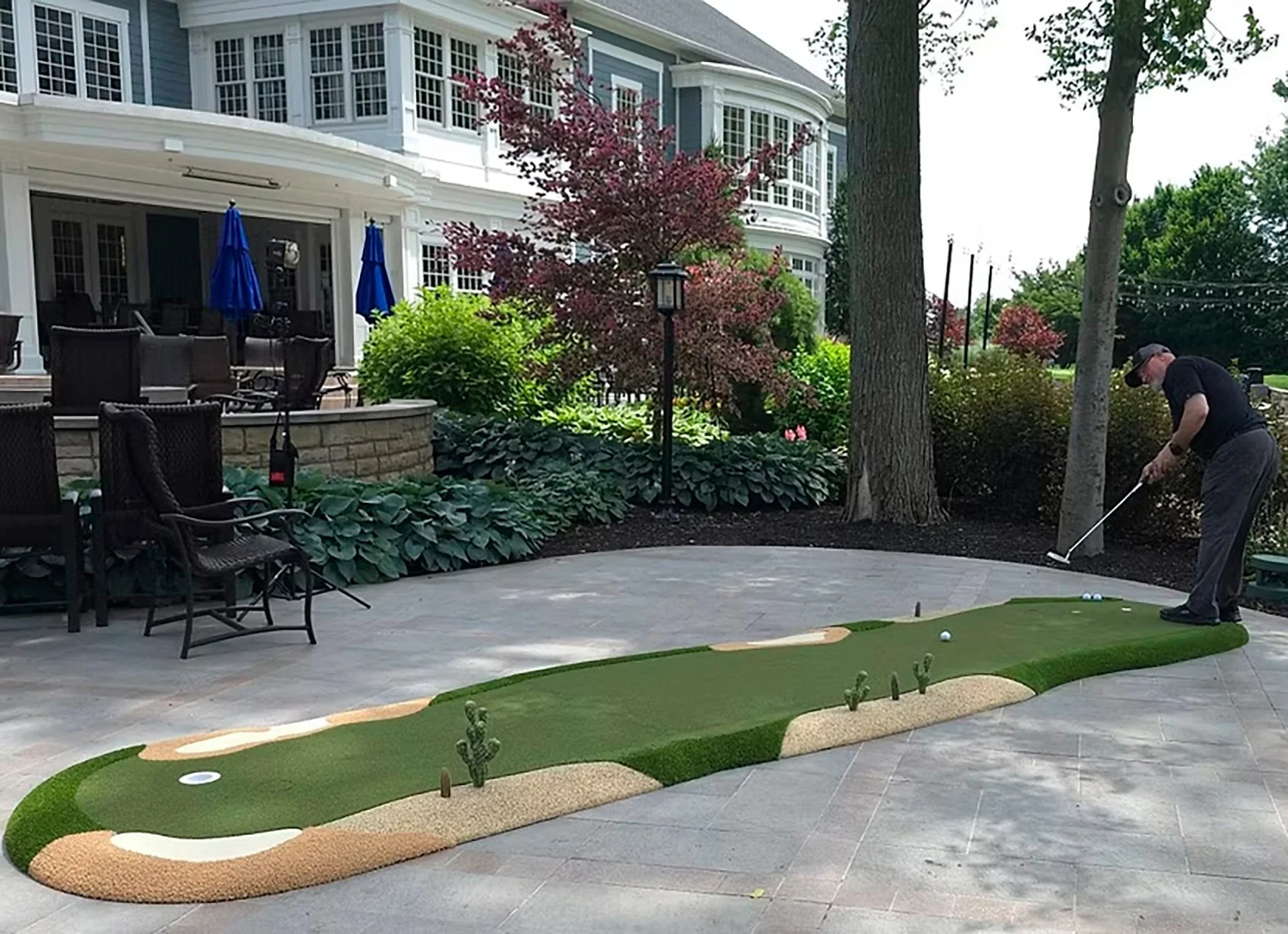 Person playing mini golf on a backyard patio with a house in the background, trees, and outdoor furniture.