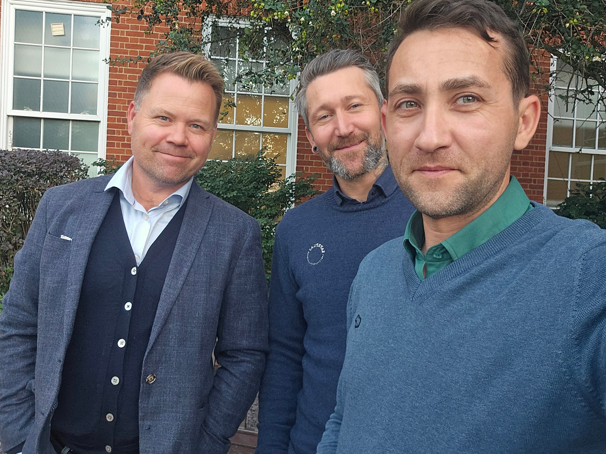 Three men taking a selfie outdoors in front of a brick building with windows and greenery.