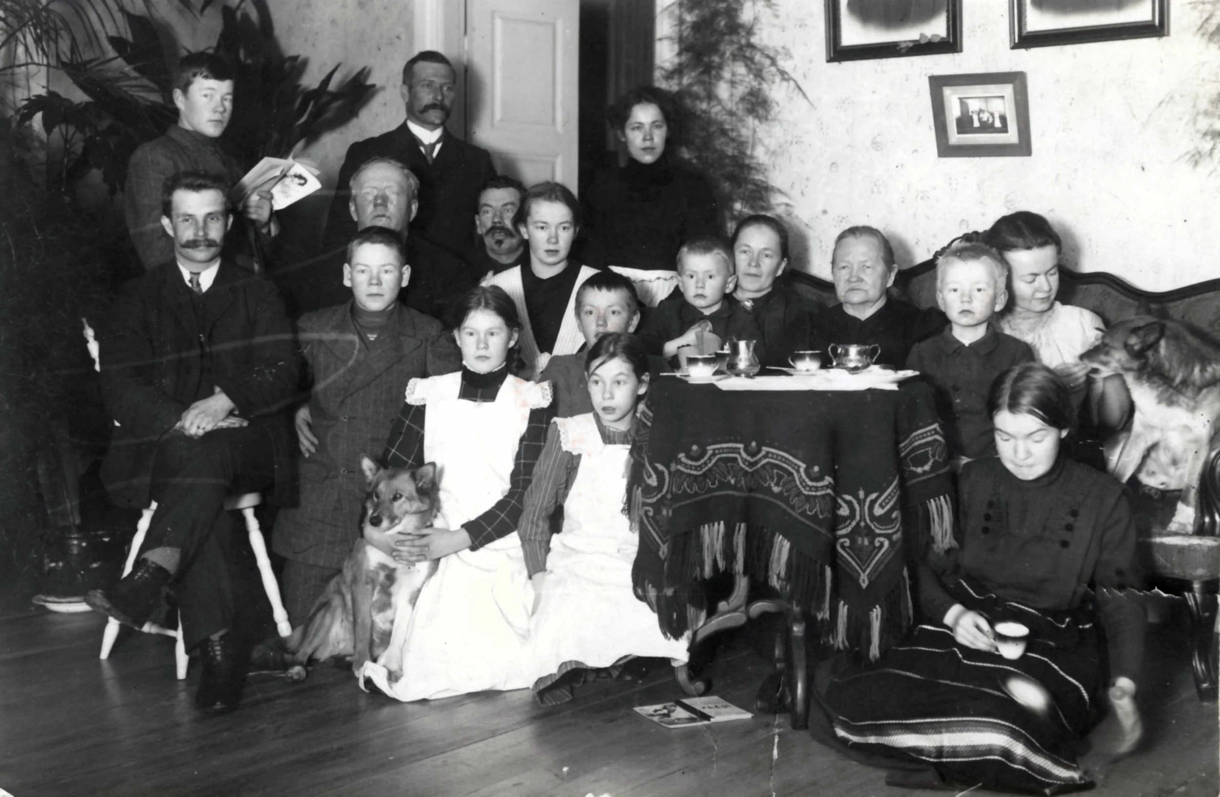 Mustavalkoinen valokuva perheestä sisätiloissa, useita aikuisia ja lapsia, joista osa istuu pöydän ääressä ja osa seistä taustalla, kaksi koiraa ja valokuvausasetelma vintage-tyyliin.