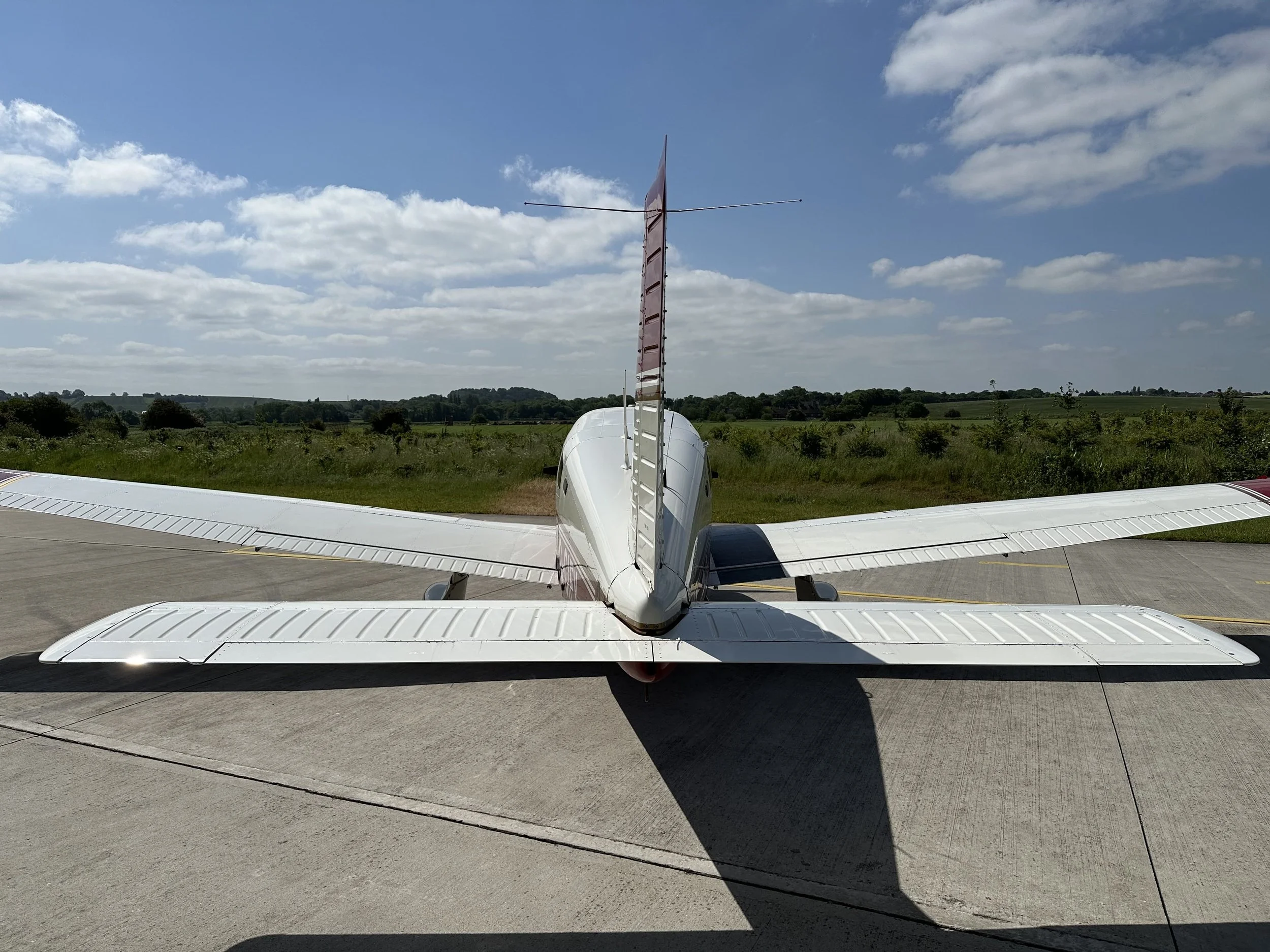 PA28 G-CIFY, Rear view of a small white aircraft with twin tail fins, parked on a runway with surrounding green landscape and a partly cloudy sky.