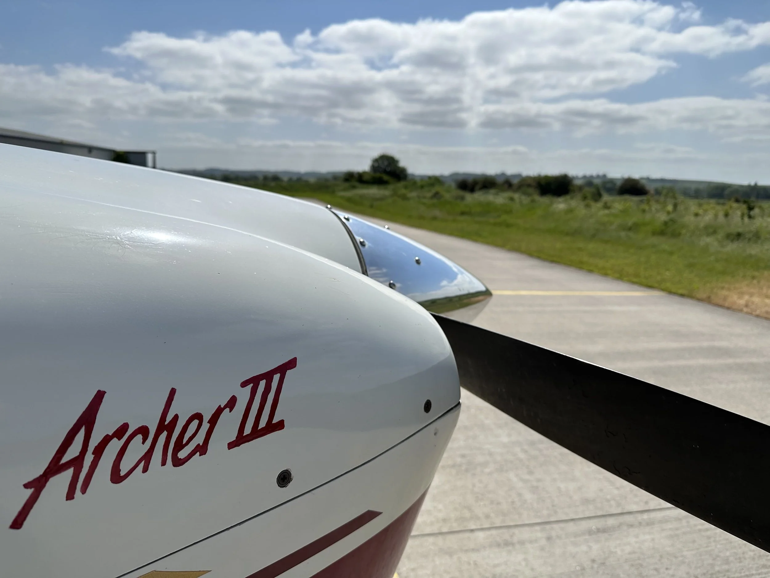 Close-up of the nose and propeller of an Archer III airplane on a runway with grass and clouds in the background.