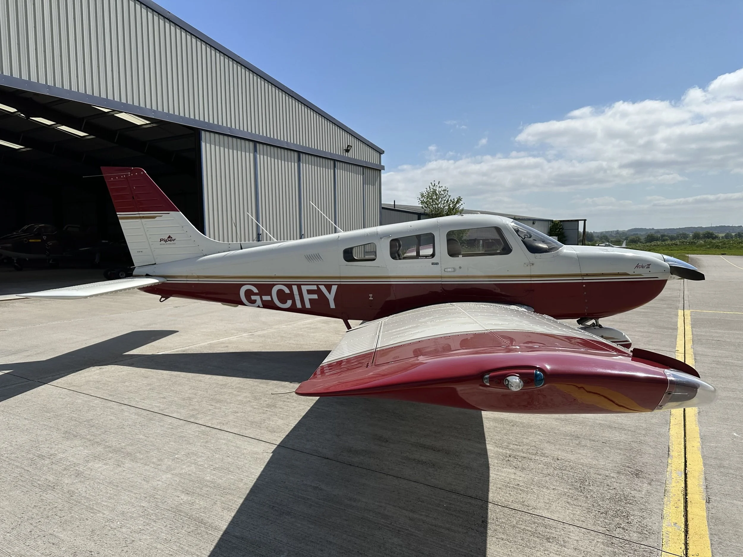 PA28 G-CIFY, Small red and white aircraft parked on an airport tarmac near a hangar under a clear sky.
