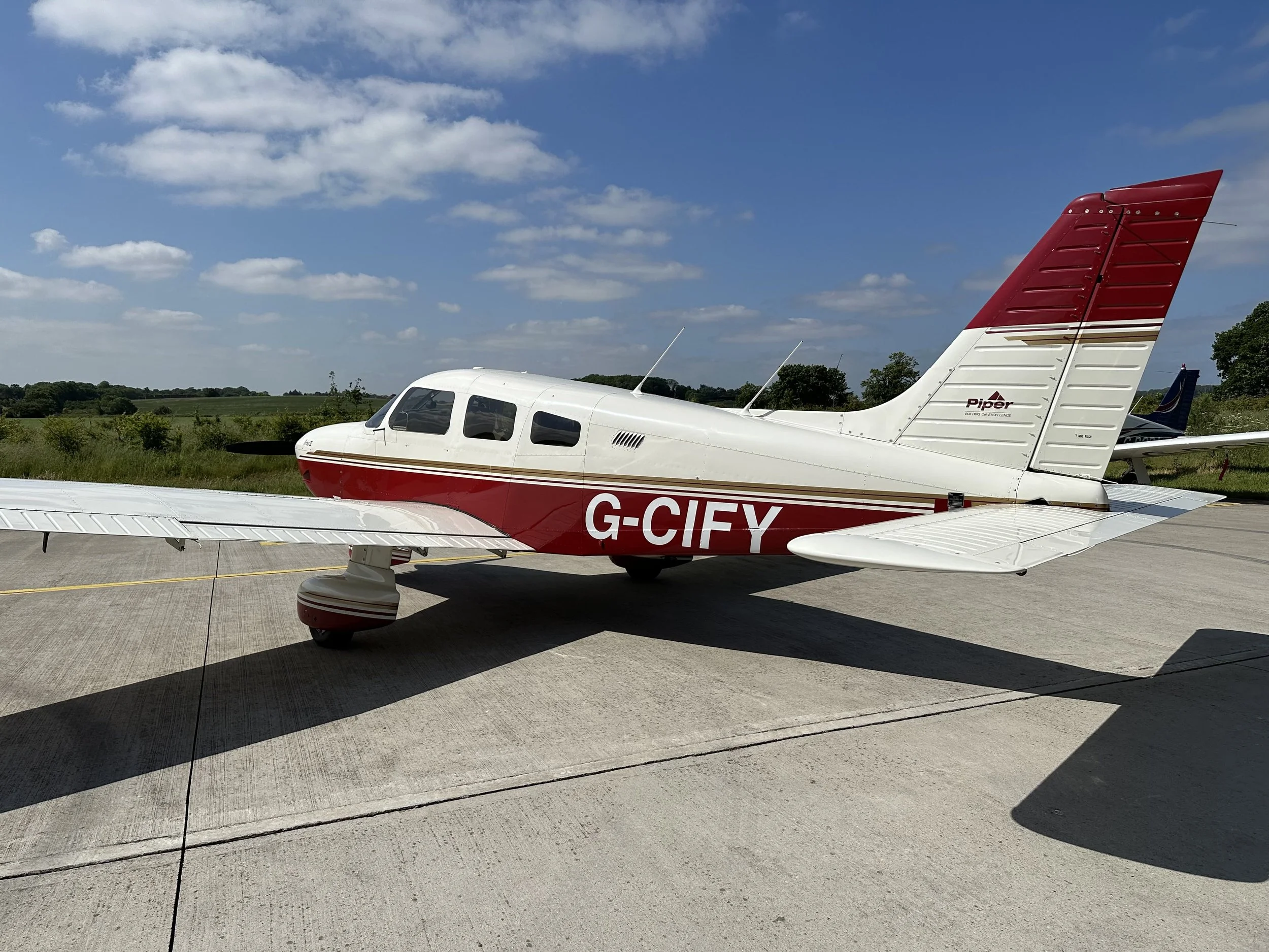 PA28 G-CIFY, Small white and red Piper aircraft parked on a runway under a blue sky with clouds.